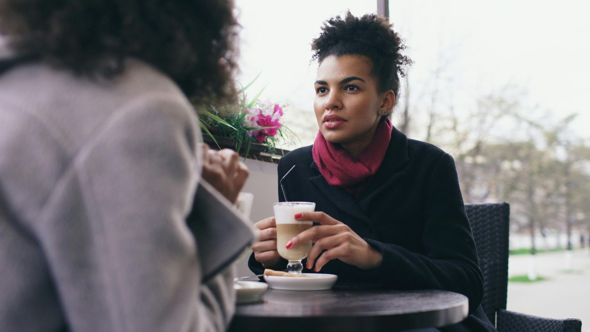 Two women having a conversation over coffee at an outdoor cafe on a bright day.