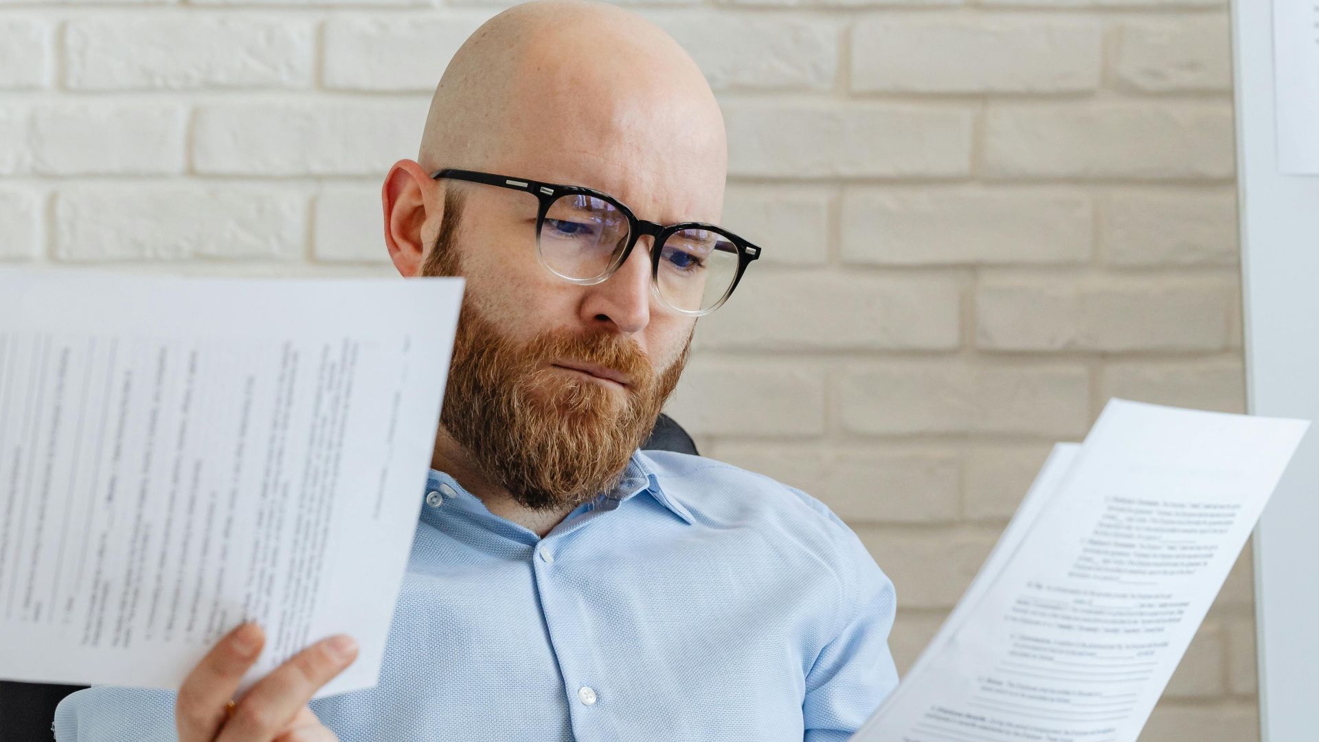 A serious-looking bald man with glasses reviews papers in an office against a brick wall.