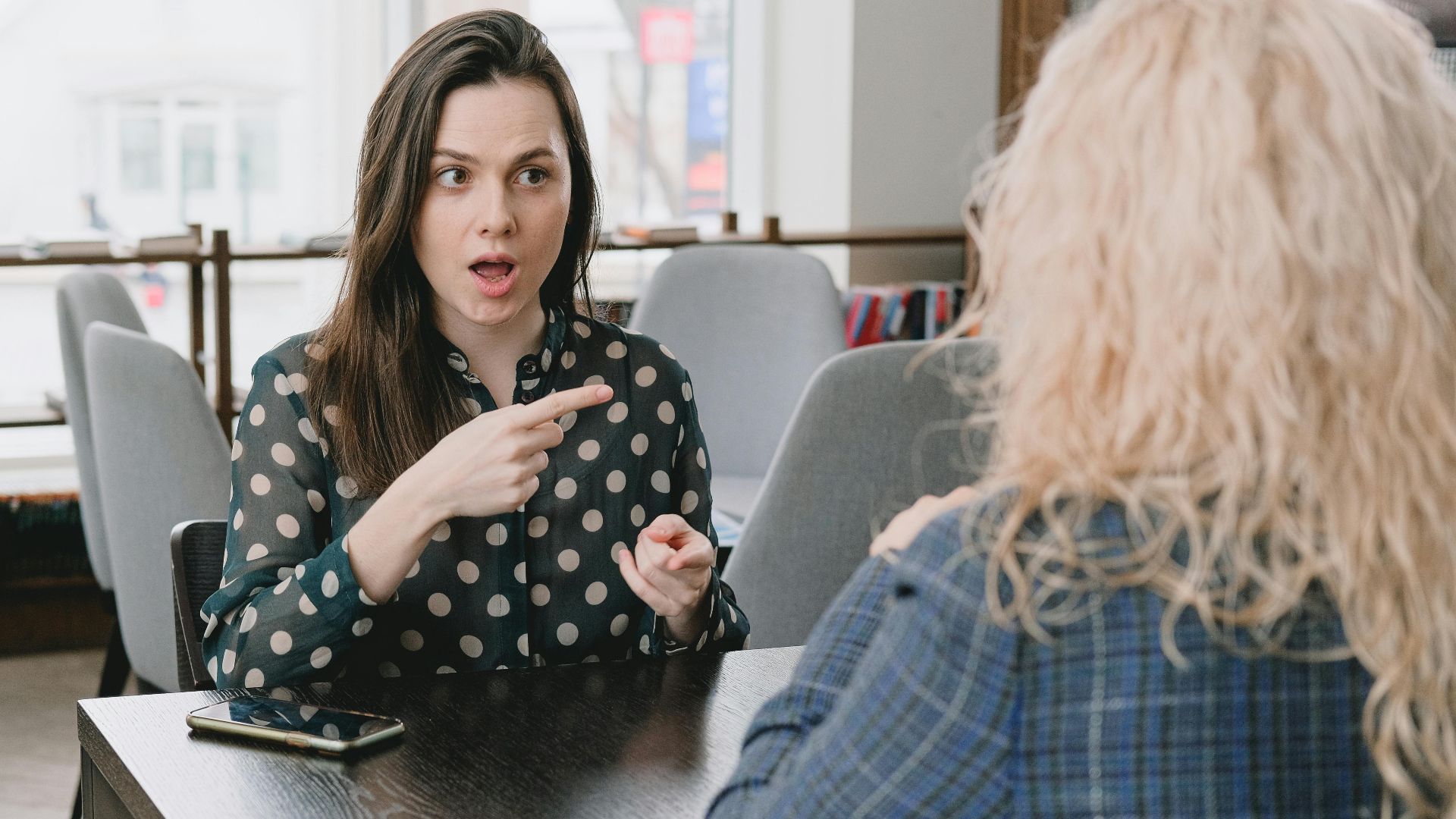 Two women in a lively discussion at a café, with expressive hand gestures and mobile devices on the table.