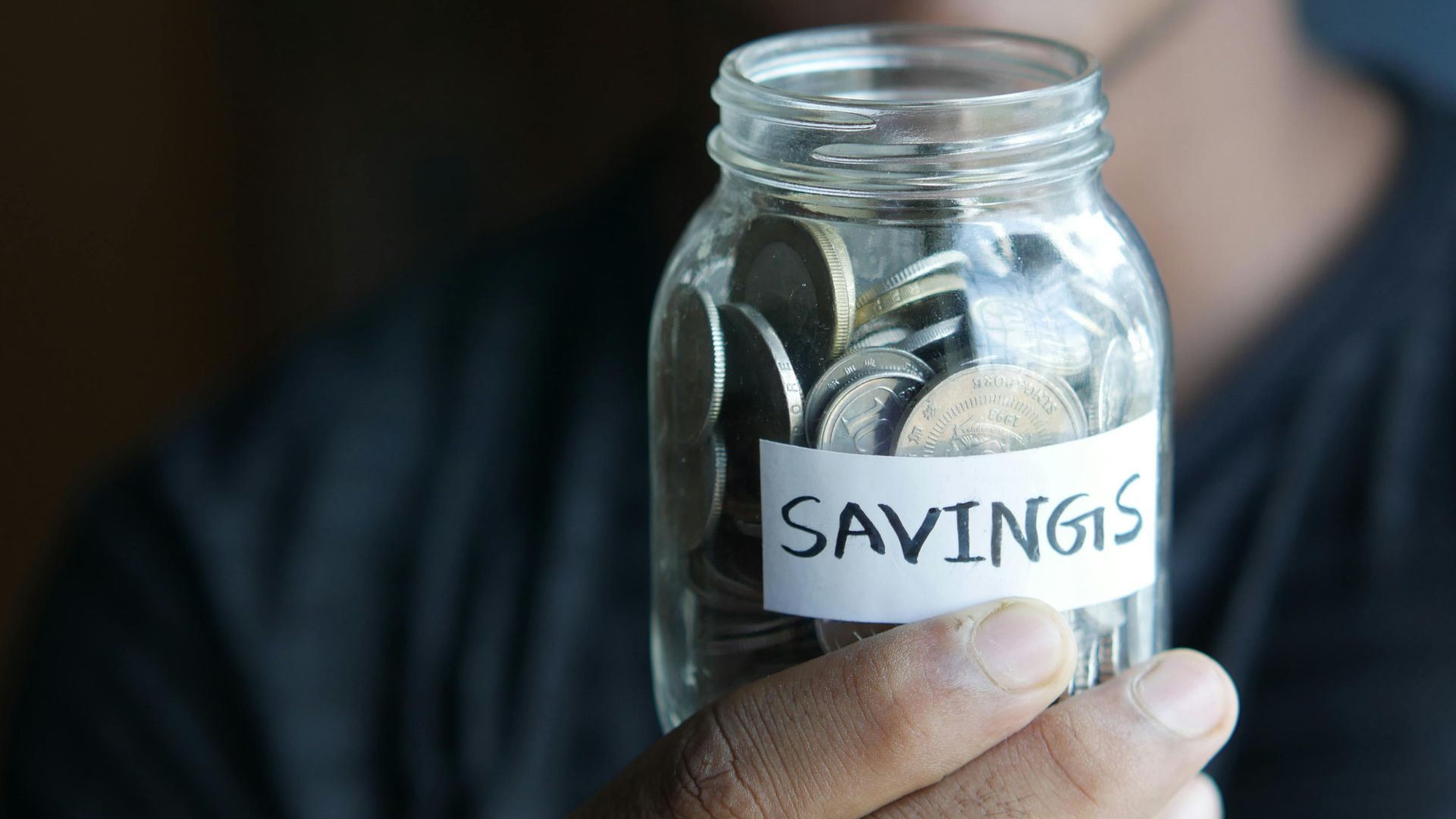 A close-up image of a person's hand holding a jar full of coins labeled 'Savings'.