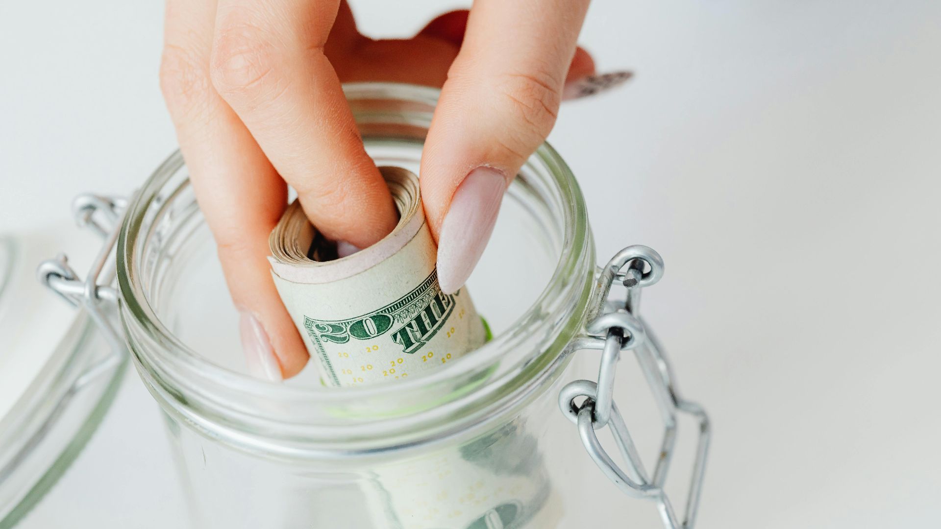 A close-up of a hand placing rolled dollars into a glass jar, symbolizing savings.