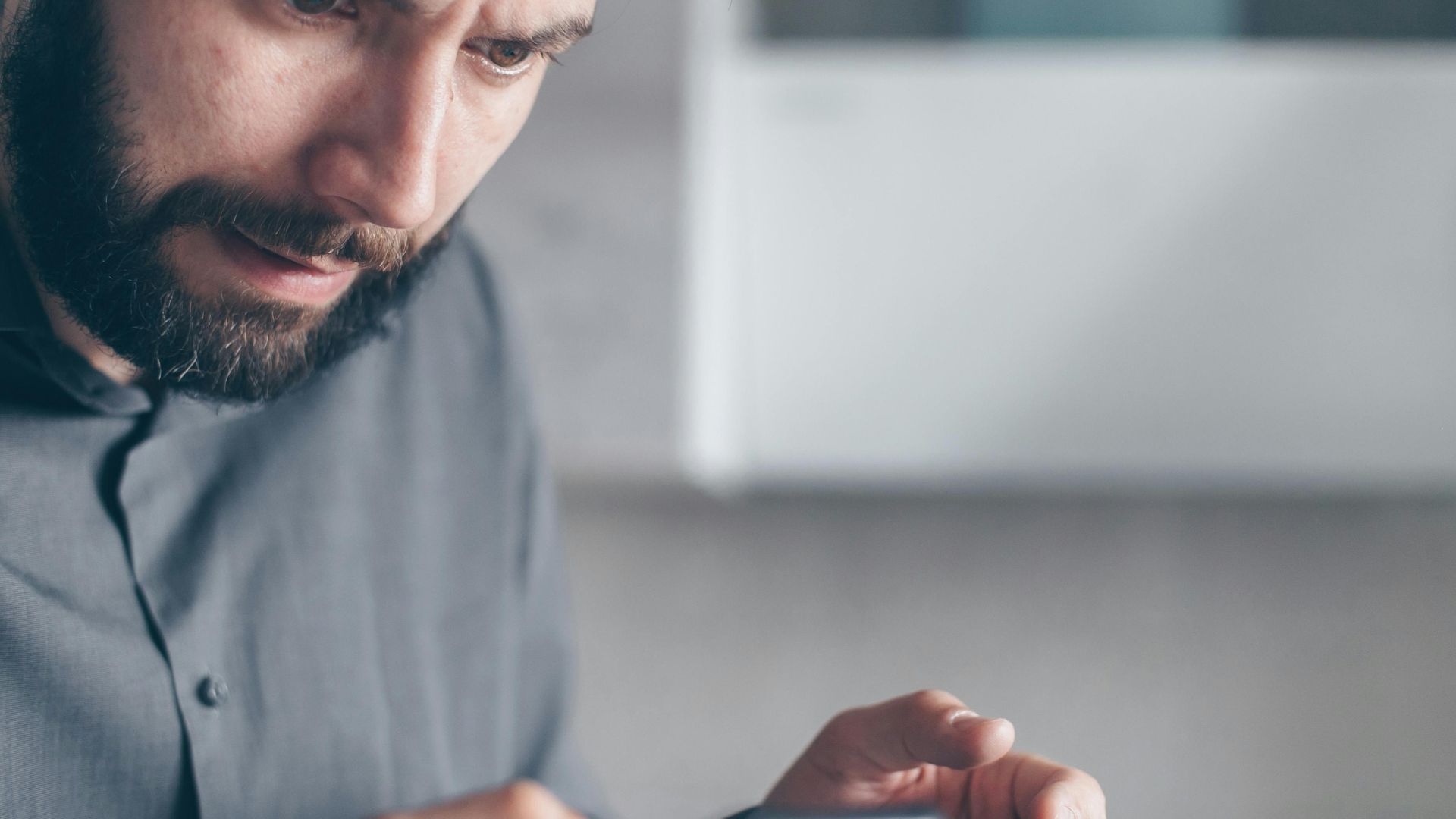 A bearded man in a gray shirt looks shocked while holding a smartphone indoors.