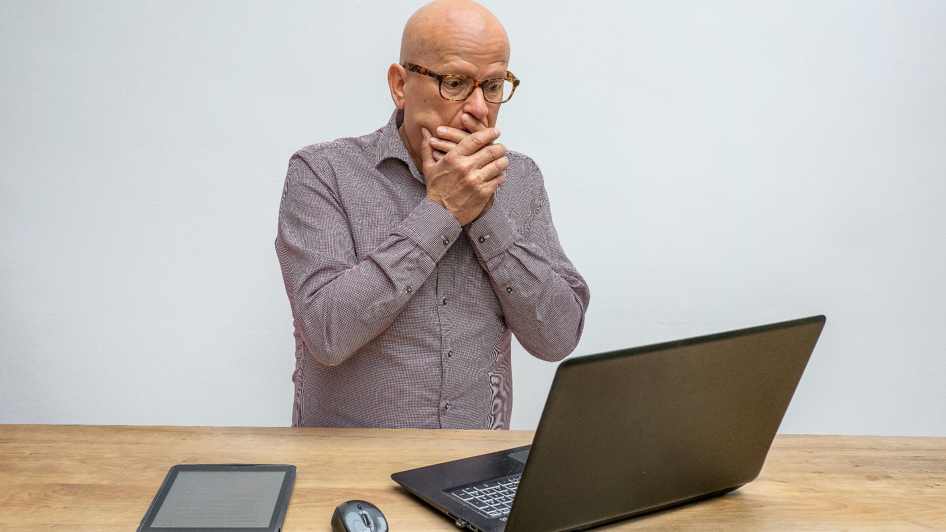 Elderly man reacts in surprise to laptop screen at wooden desk with tablet and mouse.