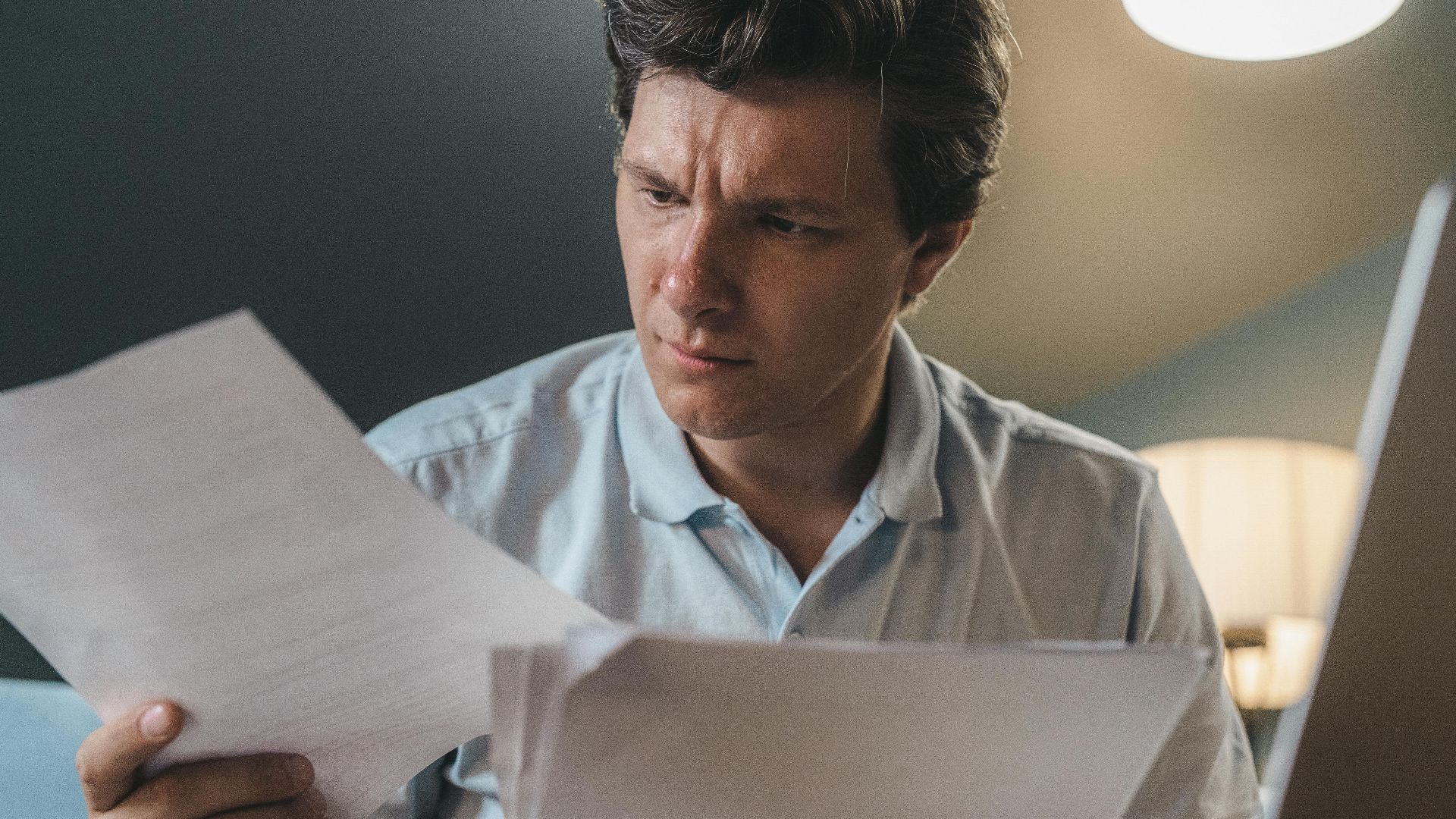 A man intently examines papers, seated indoors under warm lighting, focusing on his work.