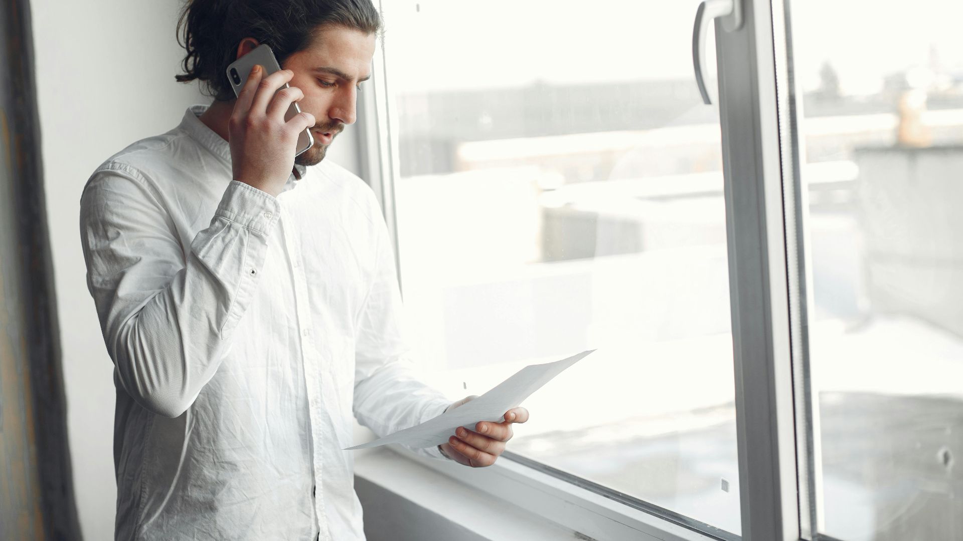 Young man in white shirt, on phone call holding a document, standing by a large window.