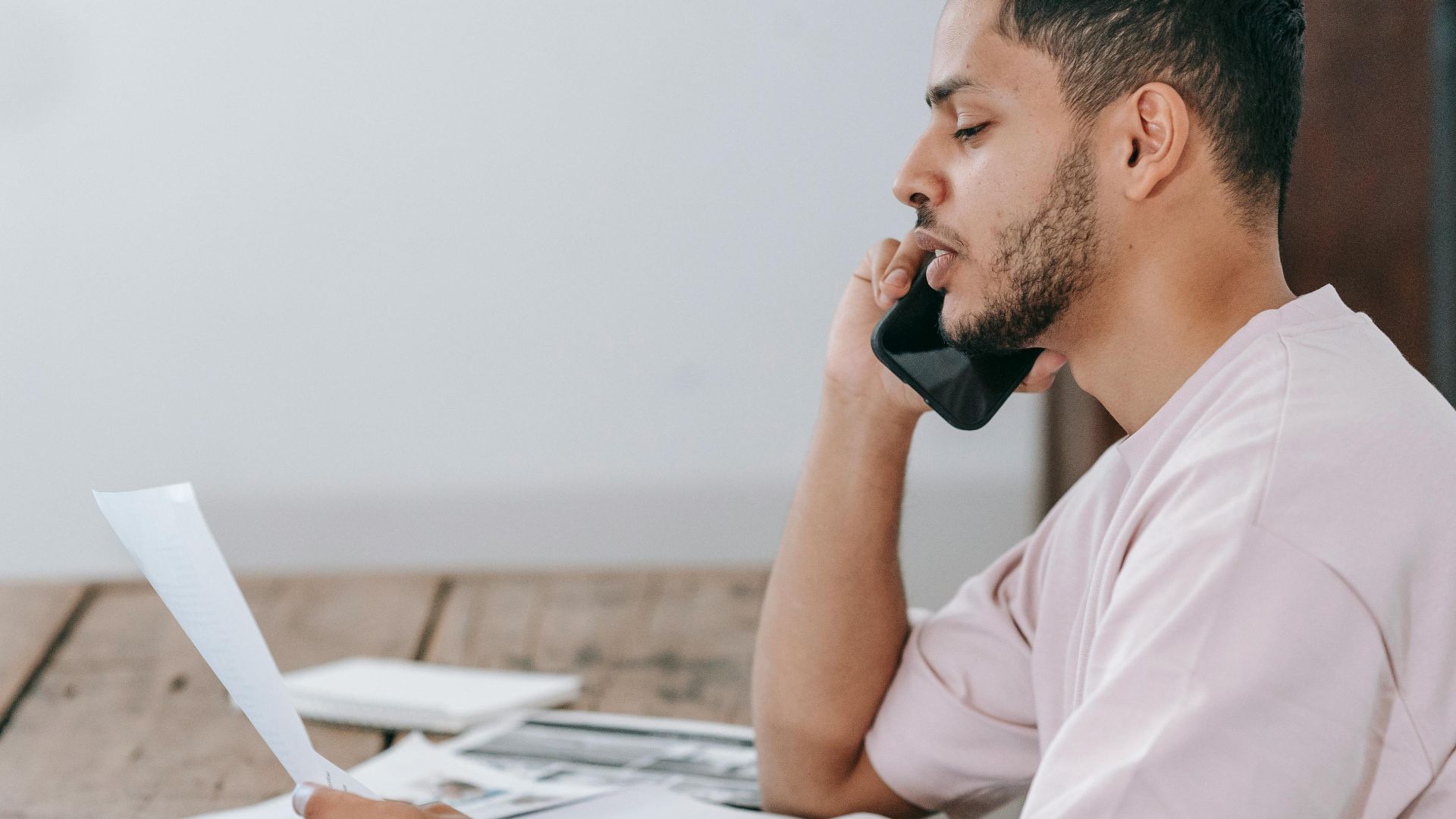Side view of young Hispanic male remote employee discussing documents during phone call while working online with laptop and papers in home office