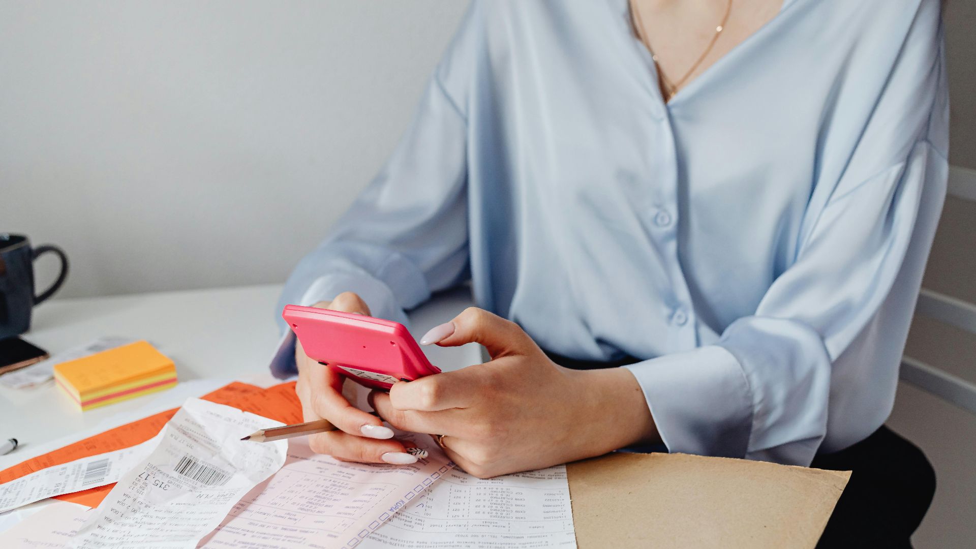 A woman using a pink calculator surrounded by bills and receipts at a desk.