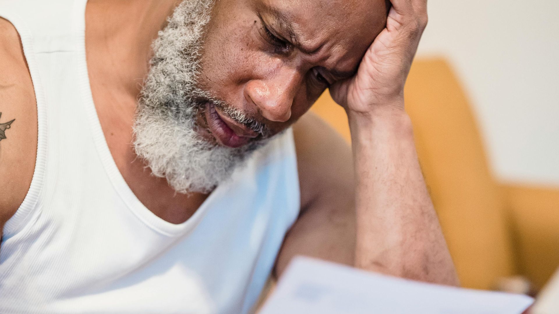 Middle-aged man in distress reading paperwork on a couch at home.