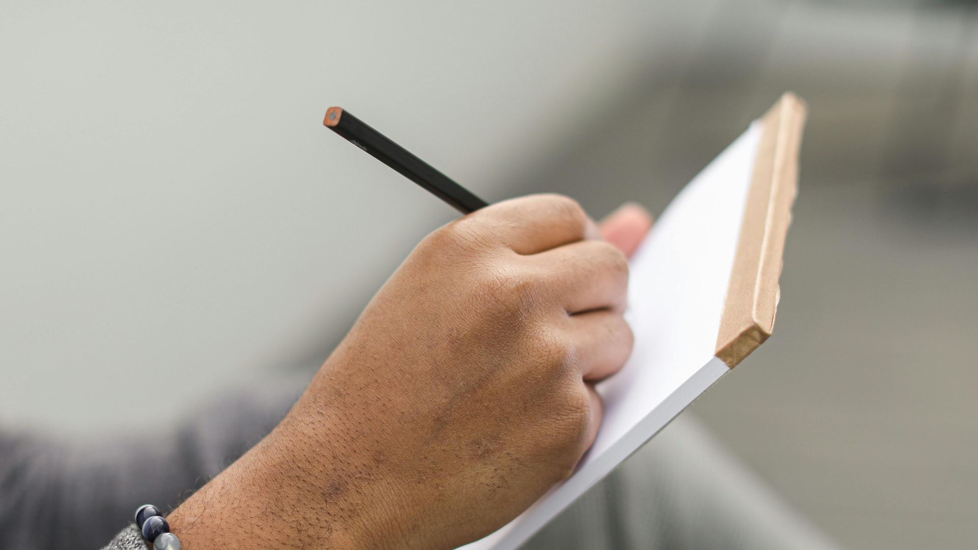 An adult hand writes in a notebook with a pencil, wearing a beaded bracelet and gray plaid sleeves.