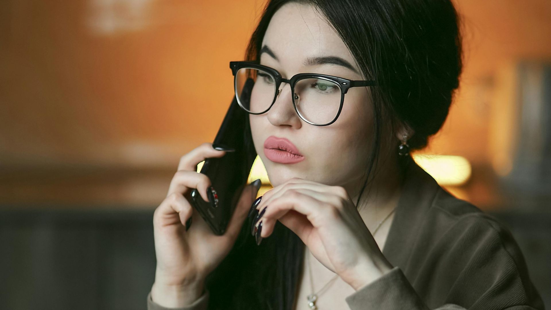 A woman with eyeglasses engaged in a phone call indoors, focused and thoughtful.
