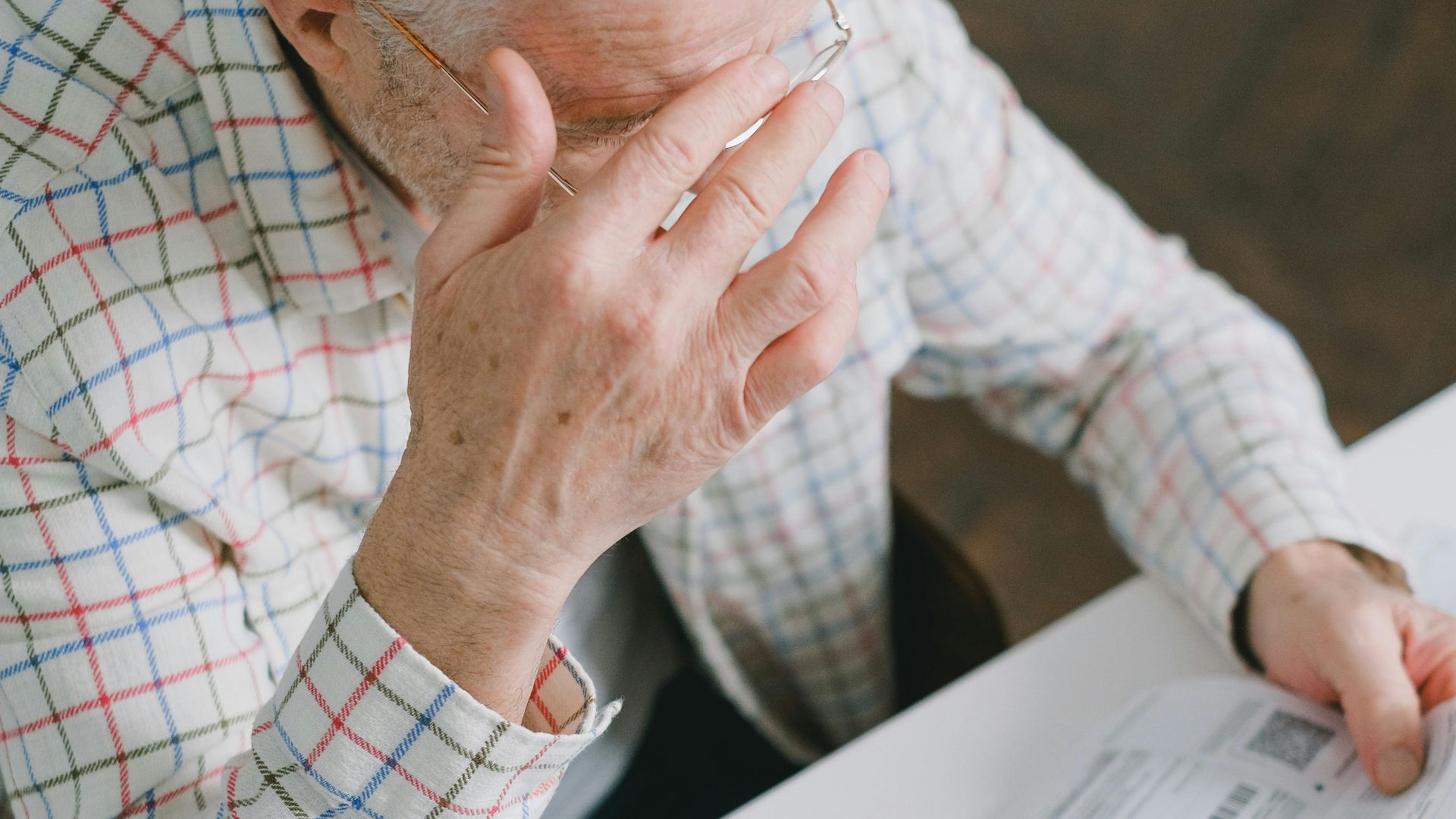 Elderly man in patterned shirt reading and holding bills at a home table, appearing focused.