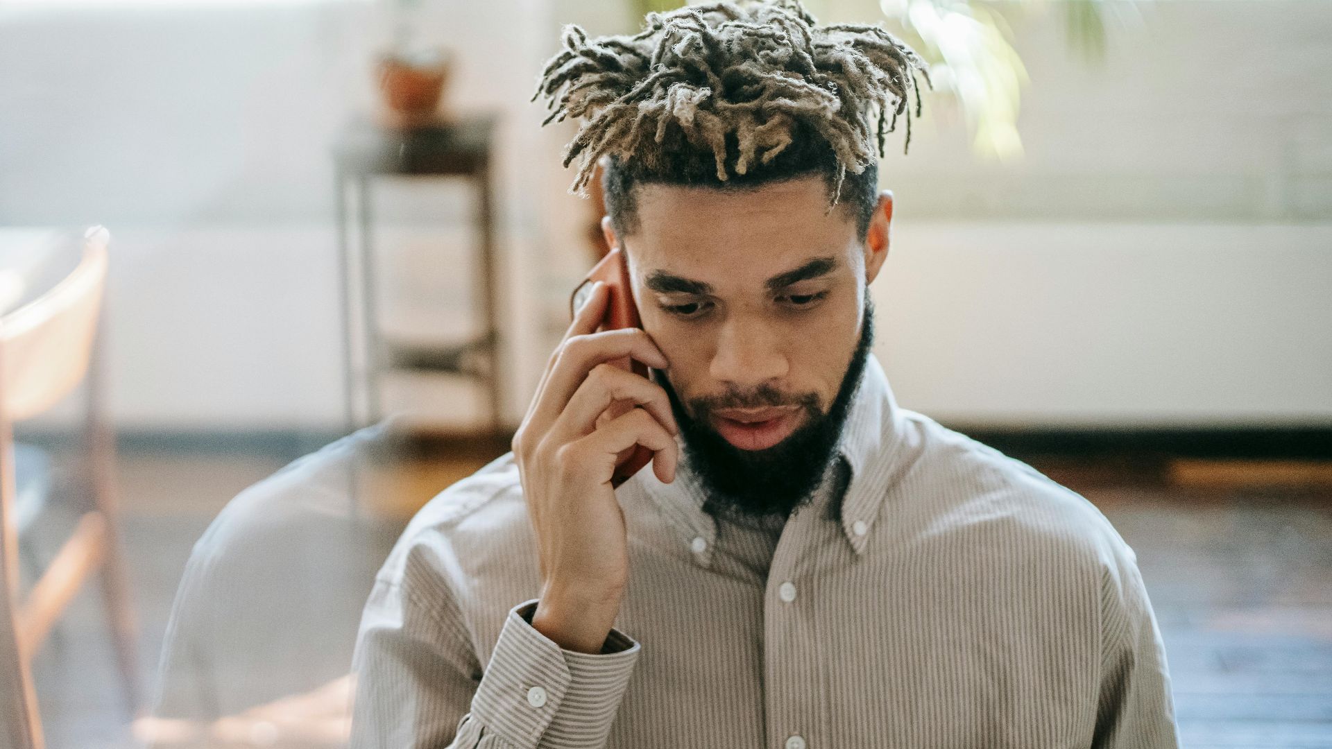Black man in casual outfit speaking on cellphone in light room near table with window and plant on background