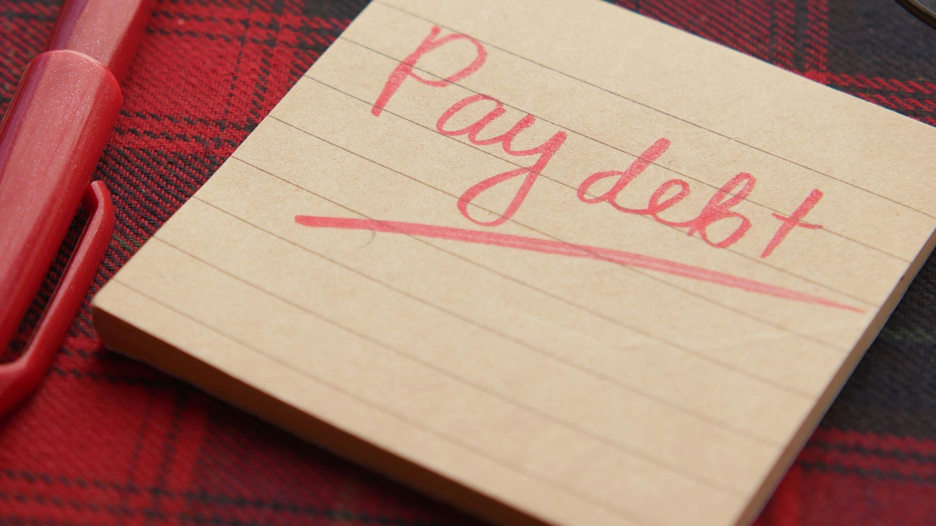Close-up of a note reading 'Pay debt' next to a red pen on a plaid fabric, emphasizing financial reminders.