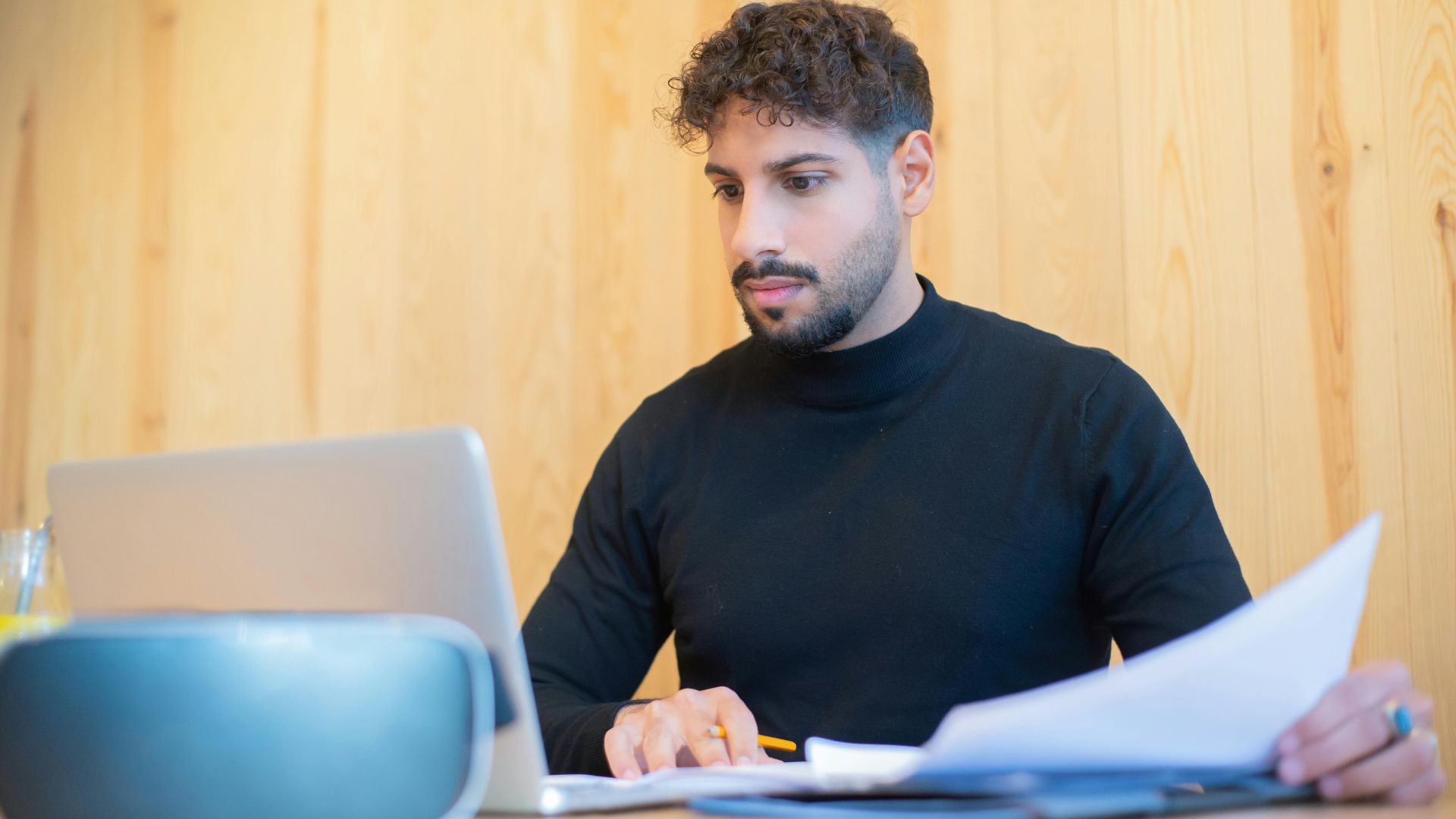 Focused businessman with beard working on a laptop, reviewing documents in a modern workspace.