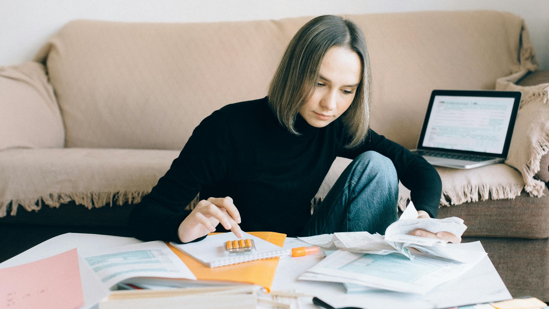 Young woman handling financial tasks with papers and laptop in cozy living room.