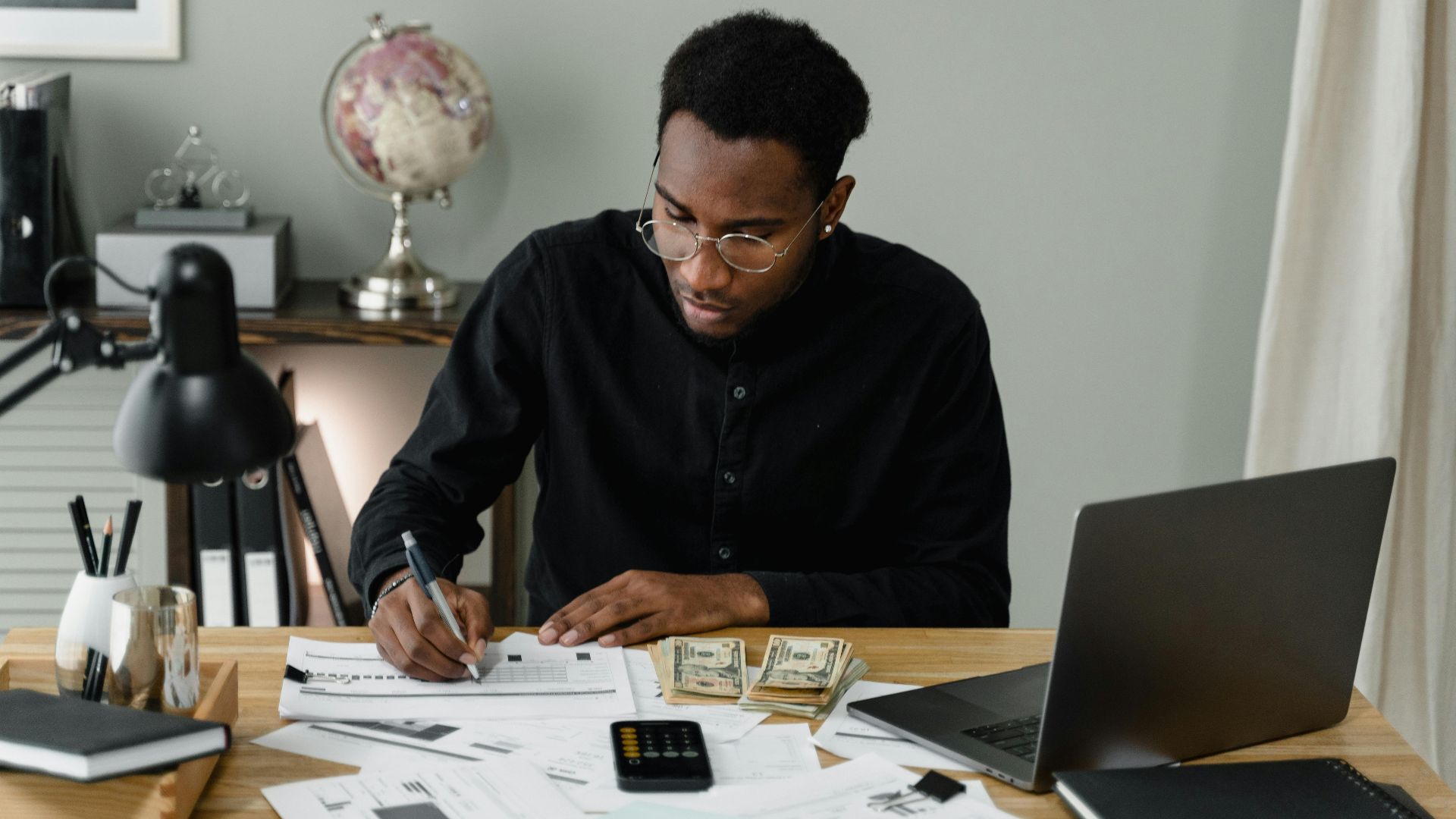 An adult man calculates expenses, using a laptop and documents at a desk in a home office setting.