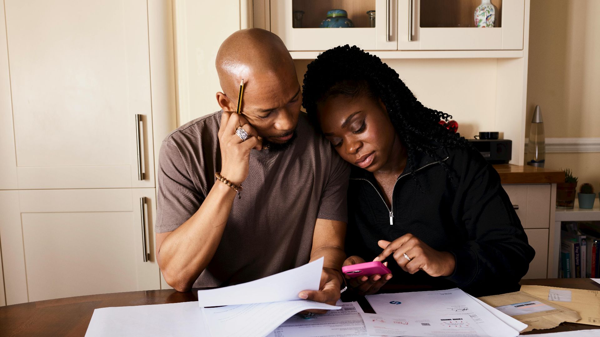 A couple sits at a table managing domestic finances, evaluating documents and using a smartphone.