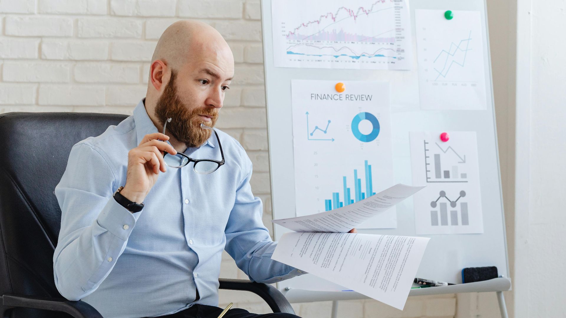 Bald businessman analyzing financial charts in modern office setting with whiteboard and paperwork.