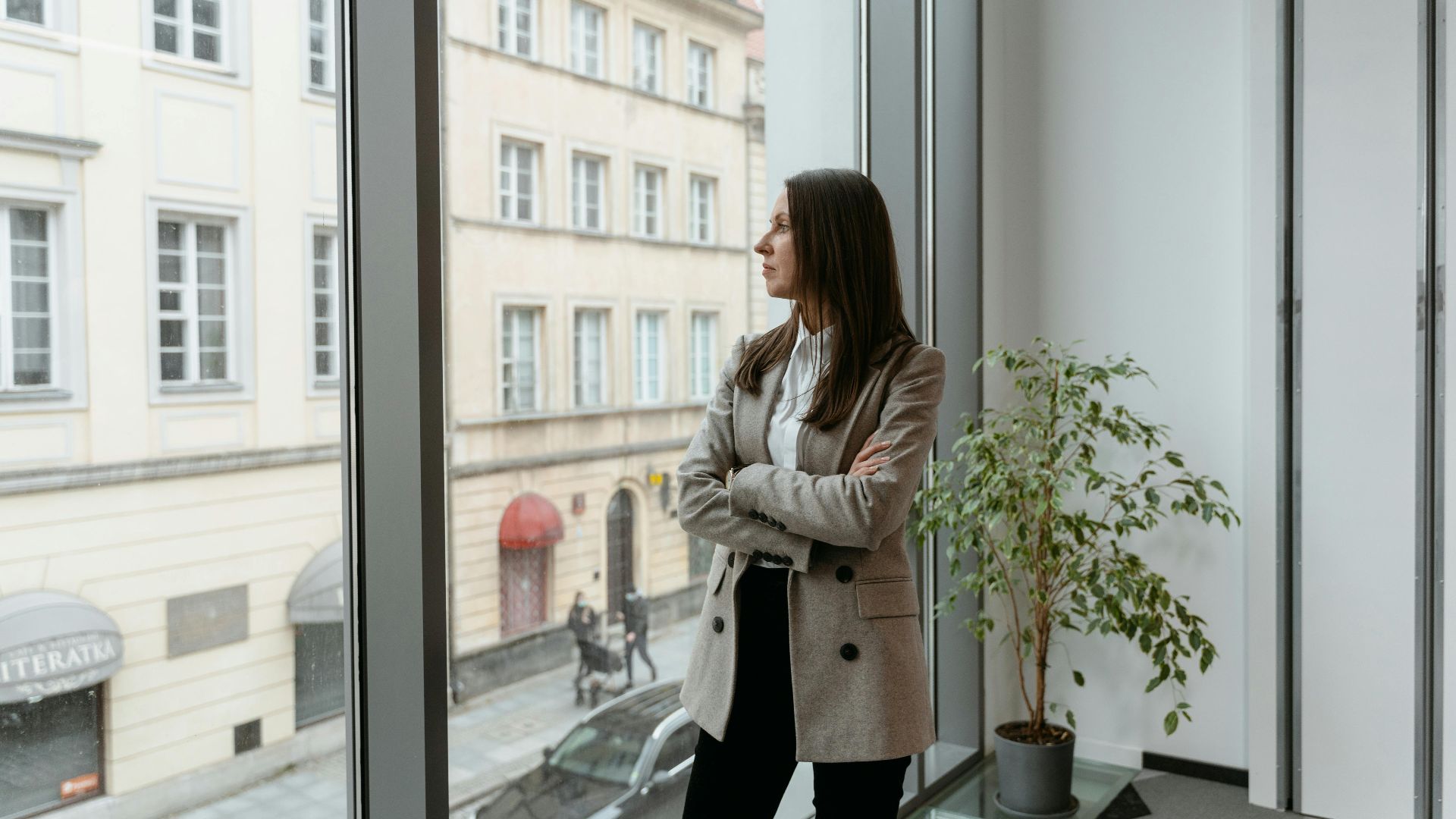A woman in a blazer stands by a large window, gazing at the cityscape from an office.