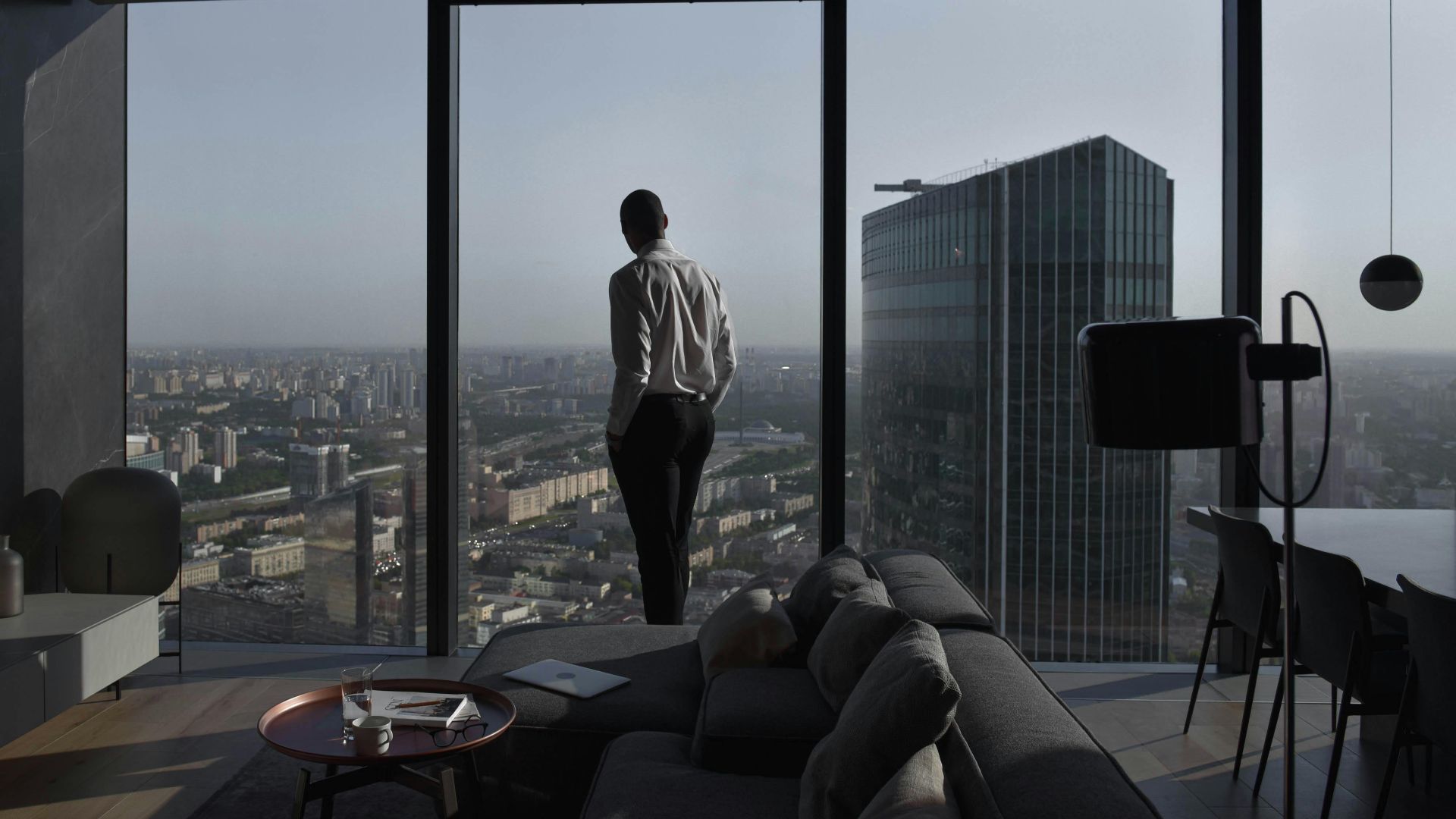Man in a modern apartment with a panoramic view of an urban skyline and skyscraper.