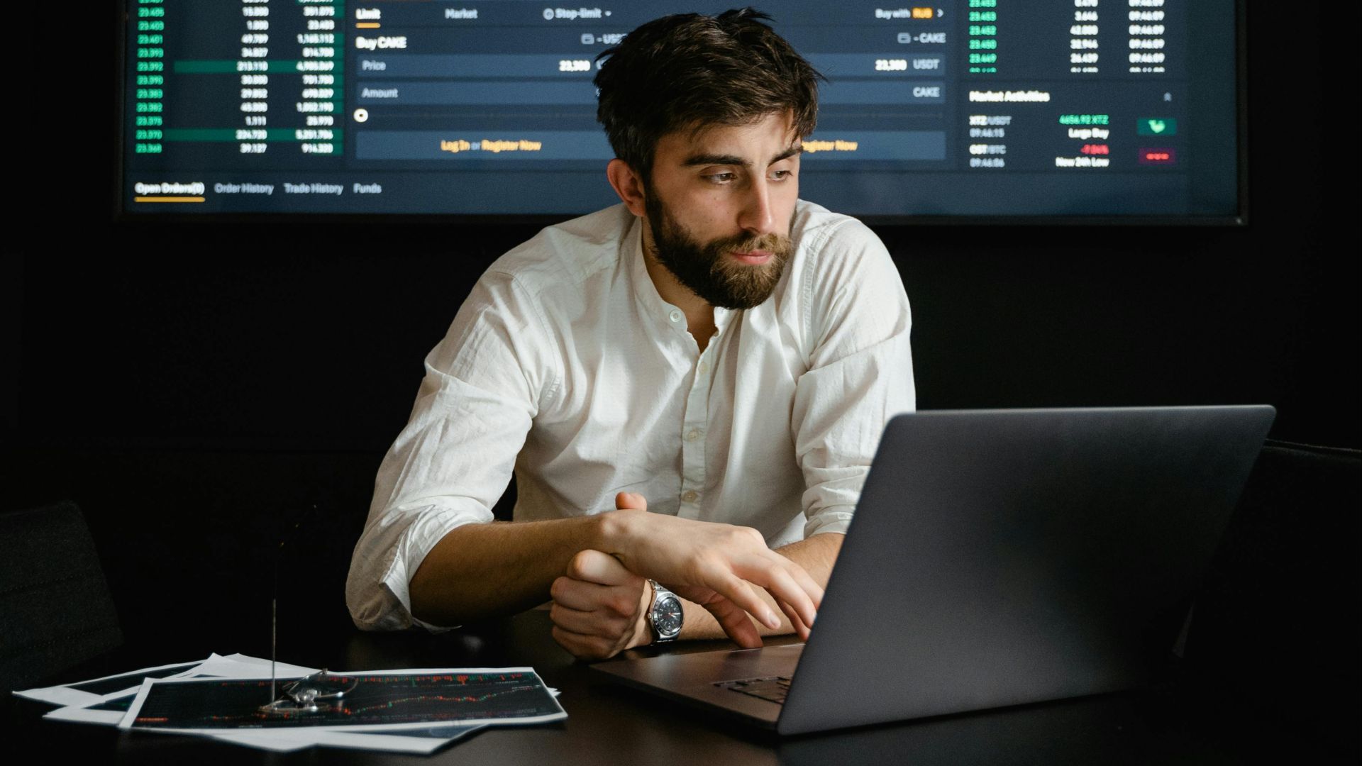 A man analyzing stock market data on a laptop in an office setting with a projector screen displaying financial charts.