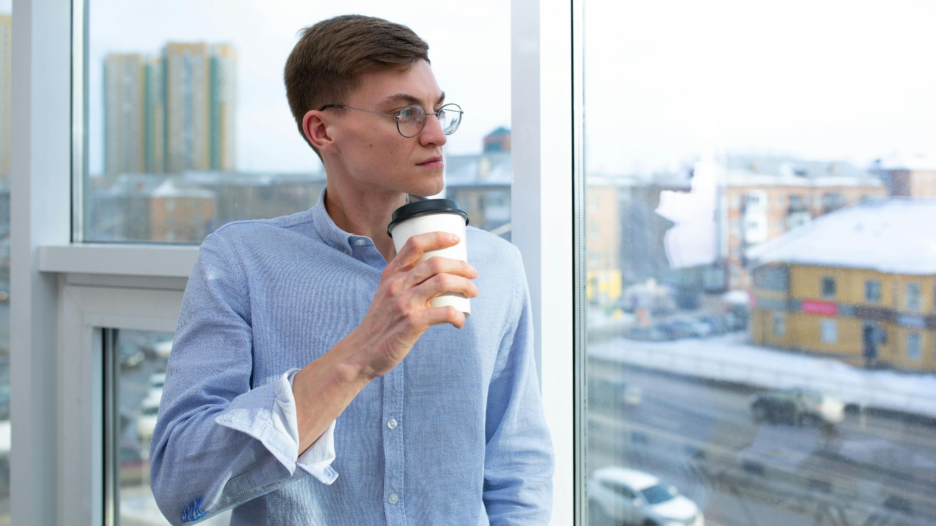 Profile of a man with glasses enjoying coffee by a large window in an urban setting.