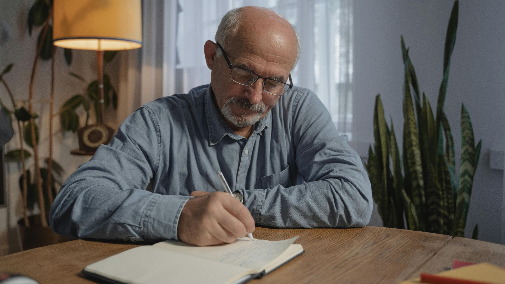 A senior adult man writing in a notebook at a desk in a cozy home office setting.