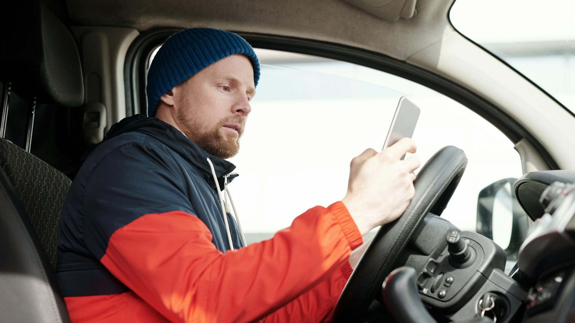 Man in a parked car using a smartphone, wearing a blue beanie and red jacket. Daytime driving scene.