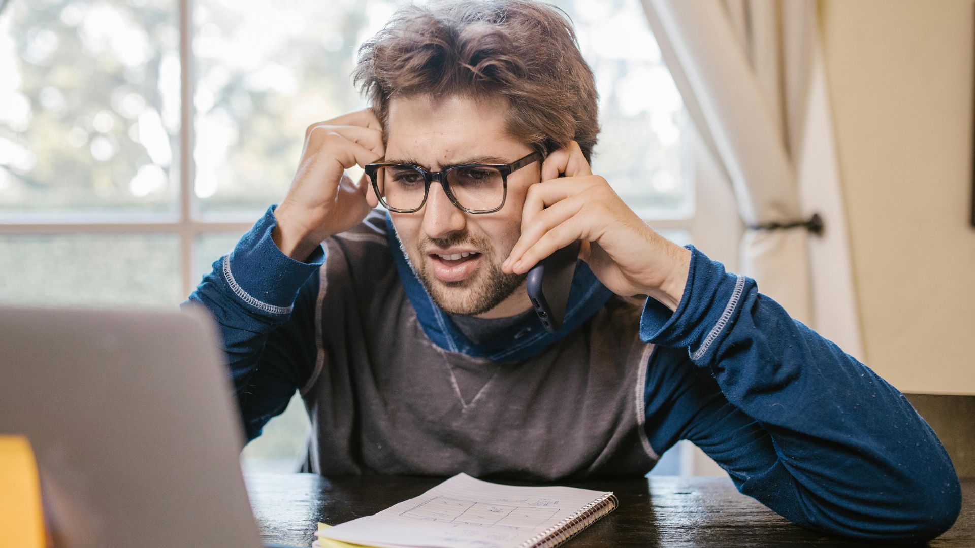 Man in glasses using laptop and smartphone at home, appearing confused.