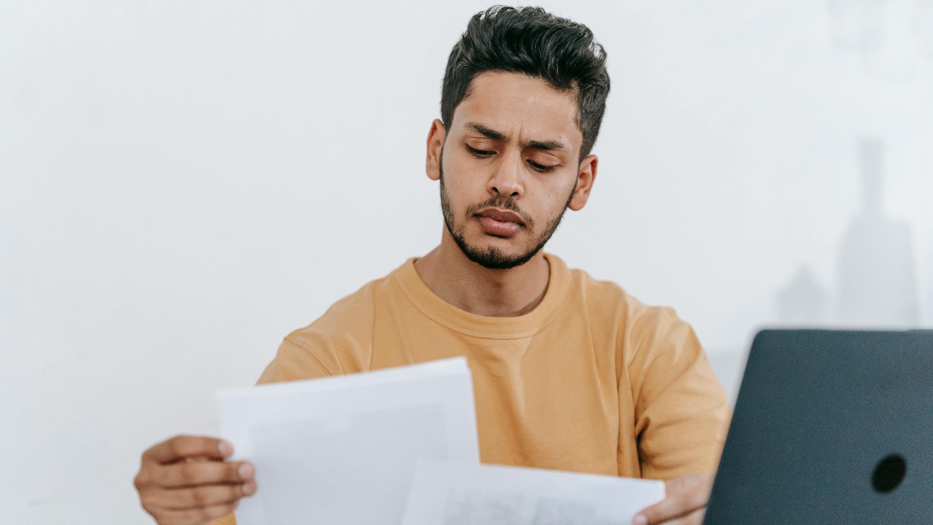 Focused young man reviewing paperwork at his desk, showcasing a business setting with a laptop indoors.