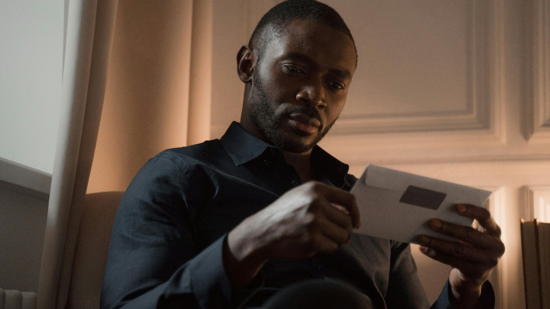 A man with a mustache reads a letter in a warmly lit room, creating a contemplative mood.