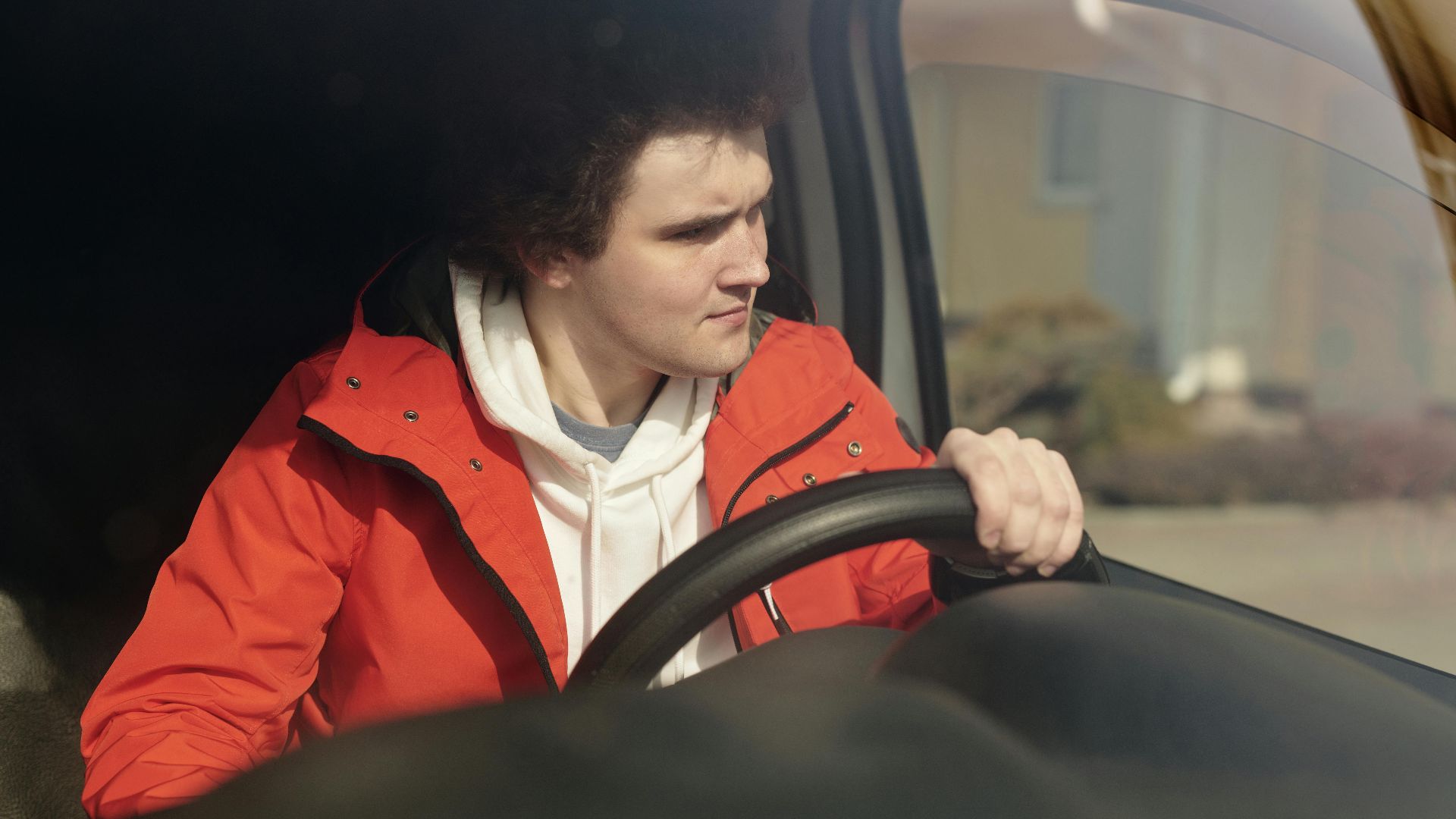 A young man in a red jacket steering a car with focus, enjoying a peaceful drive.