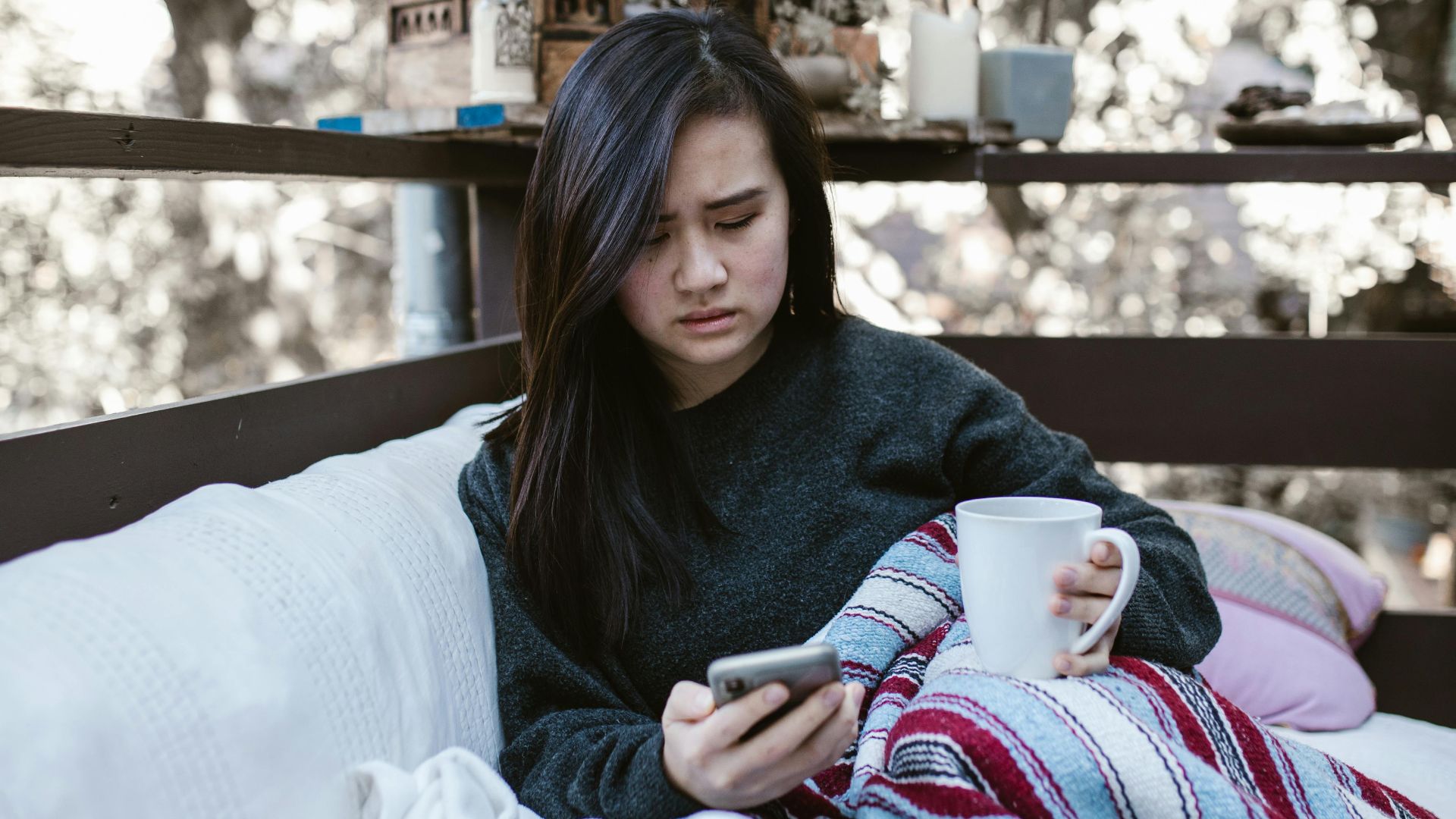 Woman wrapped in blanket, holding coffee on porch.
