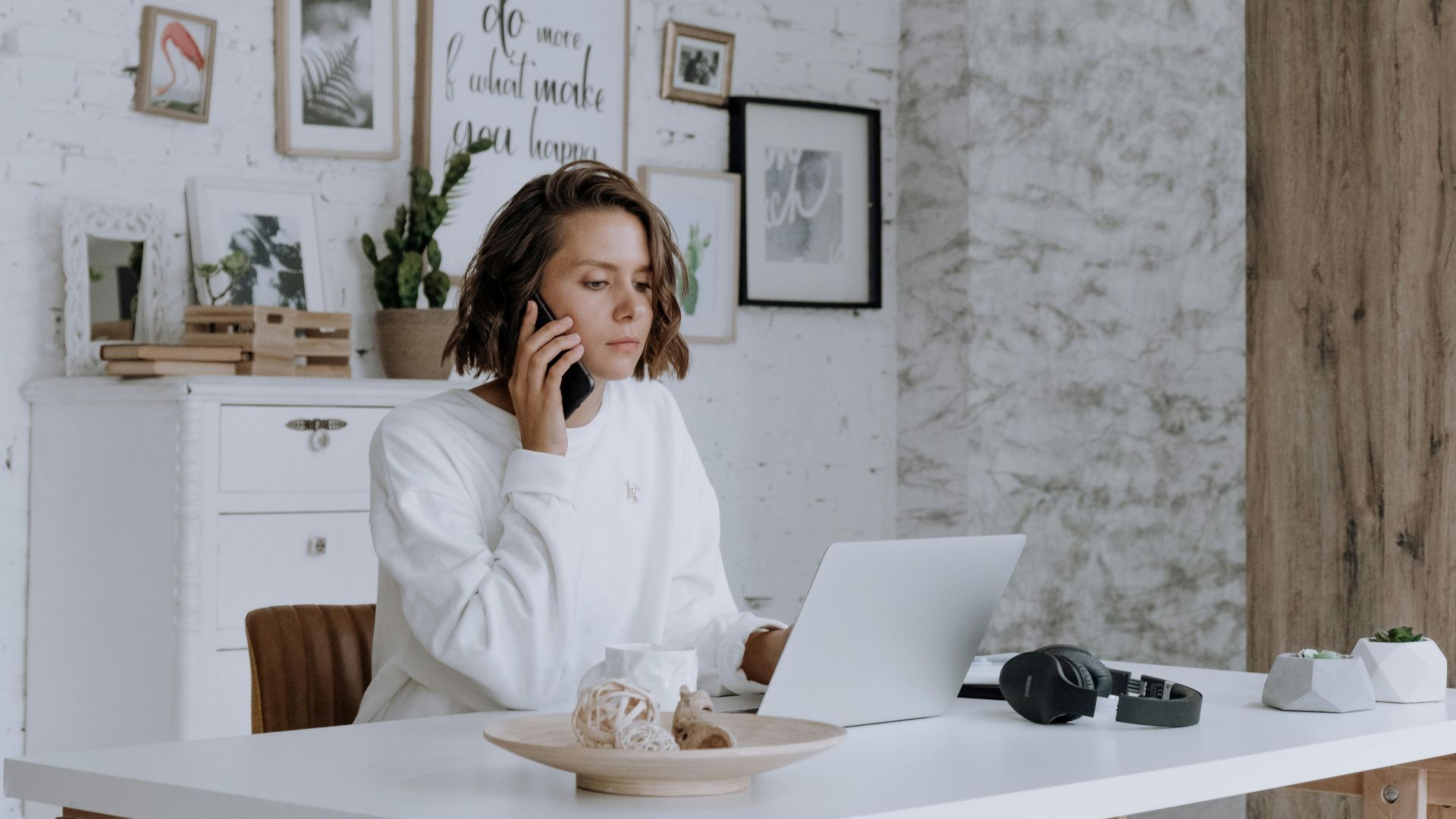 Woman talking on phone while using laptop in bright, modern home office with decor.