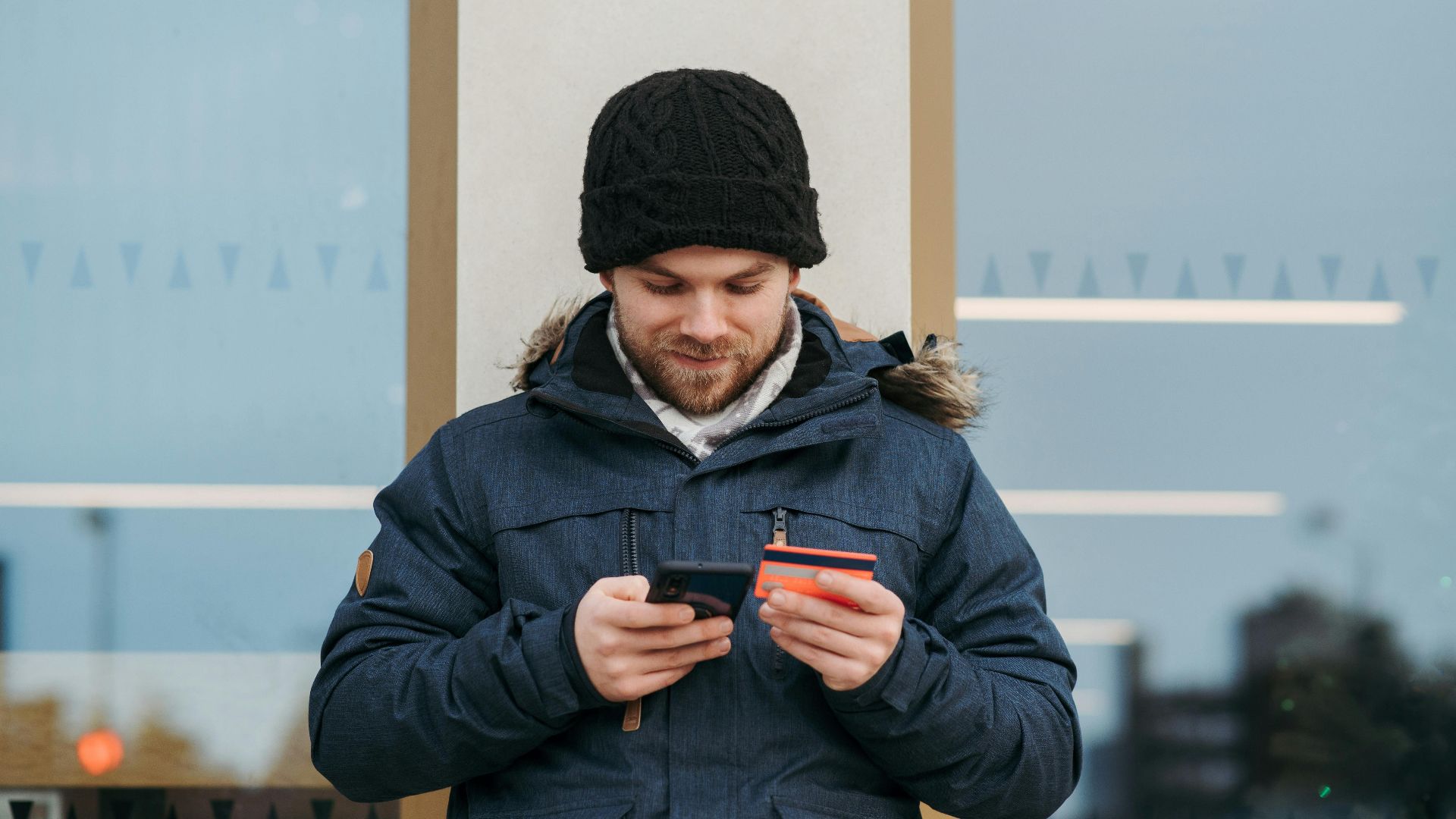 A man outdoors using a smartphone and credit card for online shopping, wearing winter clothes.