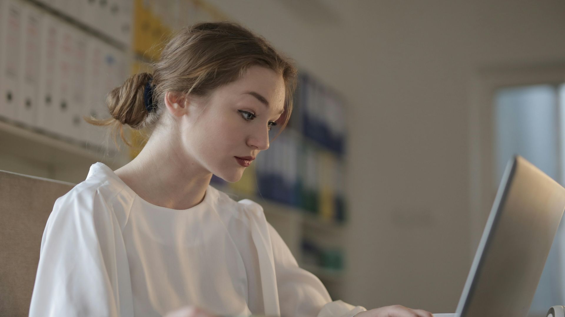 Focused woman in white working on a laptop in a modern office setting.