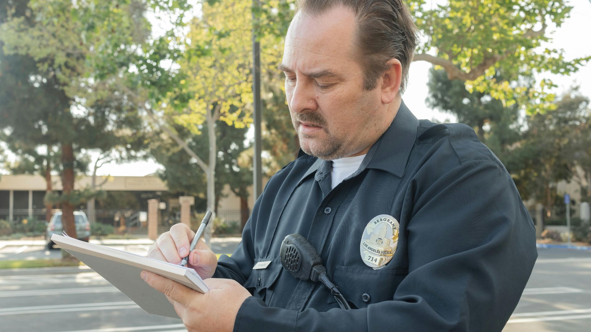 A police officer outdoors writing a report on a notepad. Trees in the background.