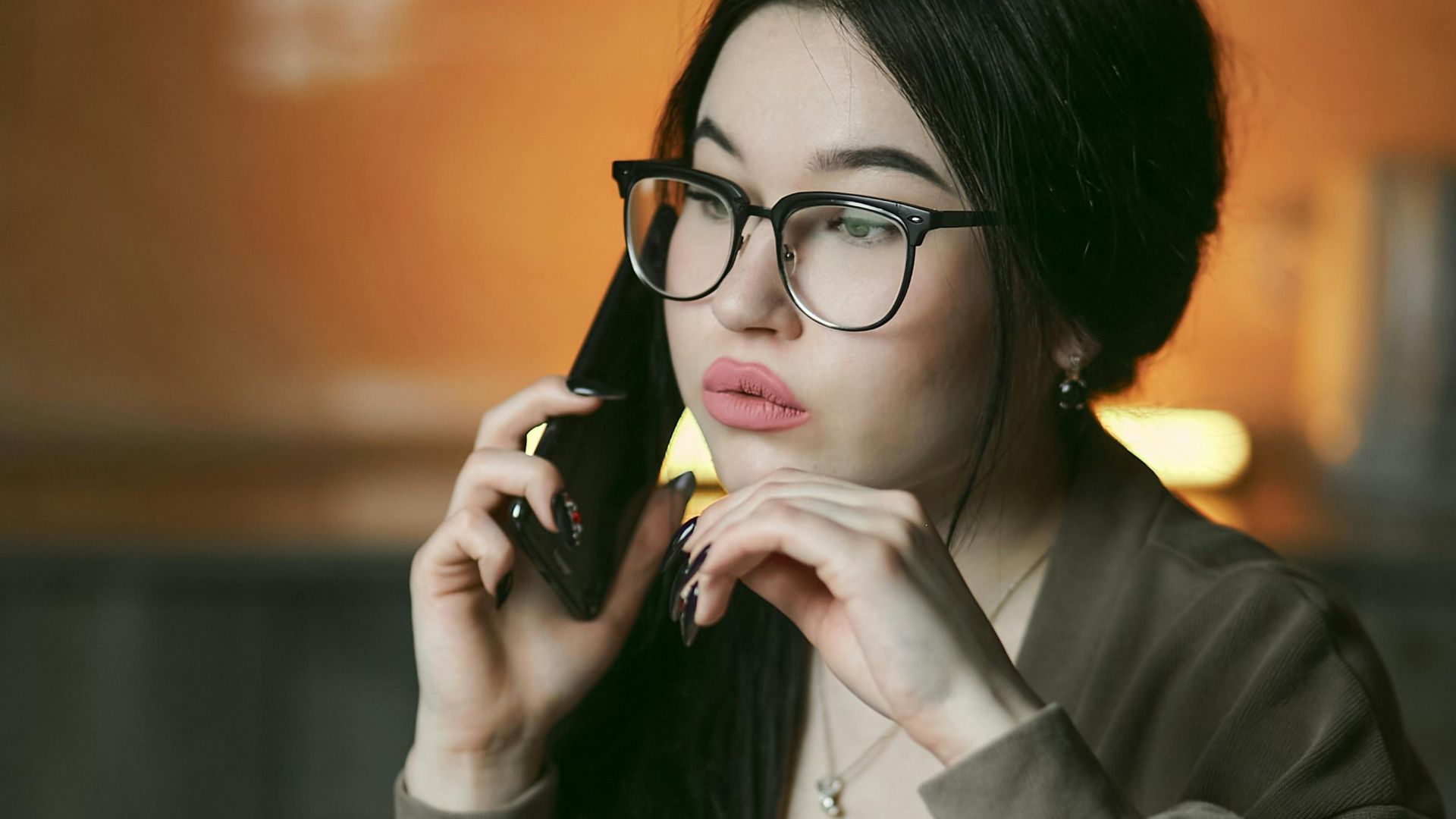 A woman with eyeglasses engaged in a phone call indoors, focused and thoughtful.