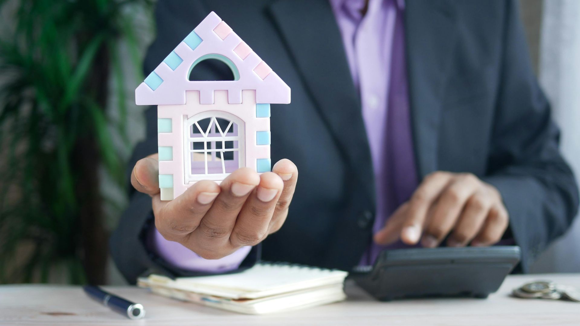 Businessman in suit holds model house, calculates real estate investments.