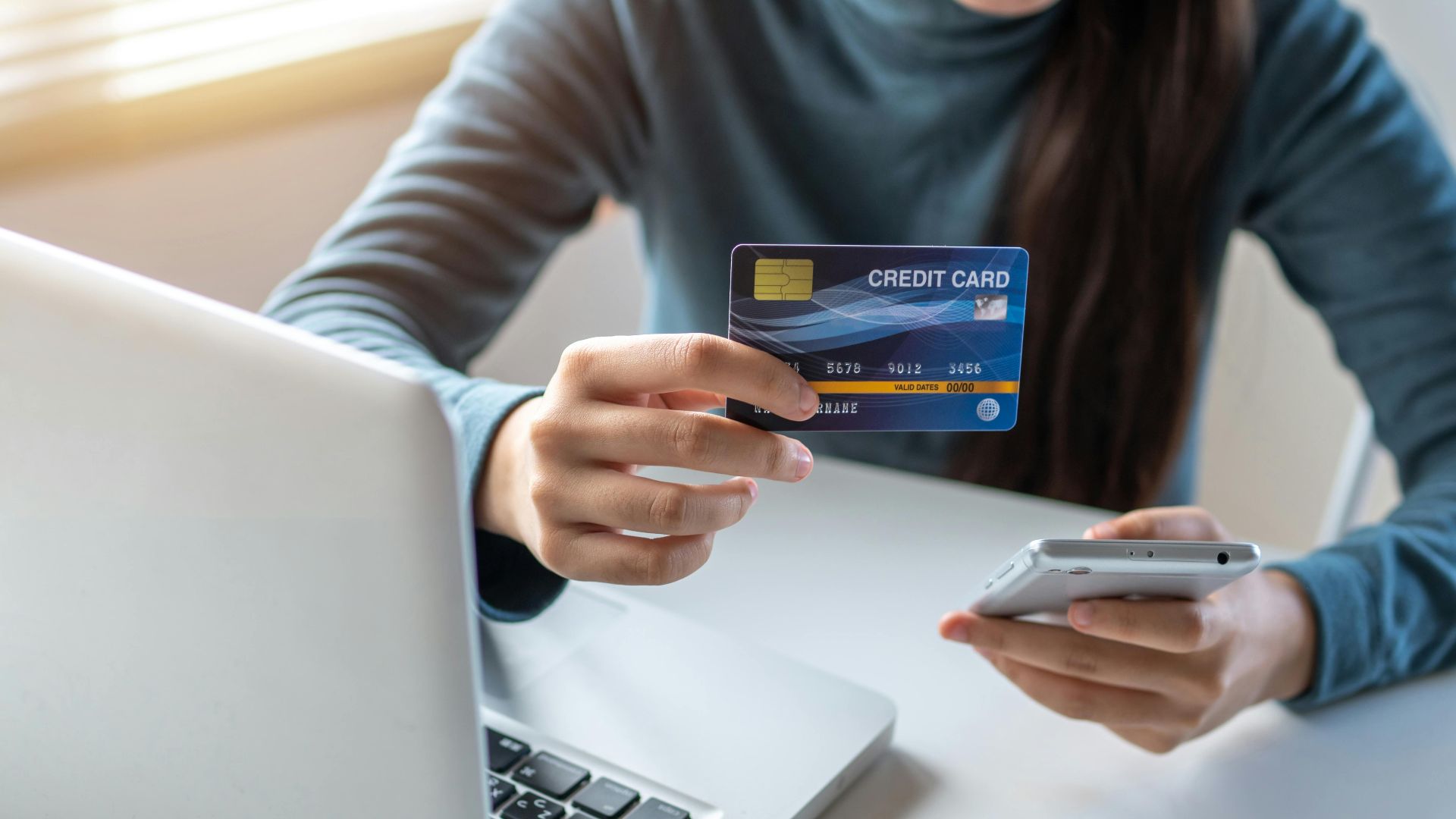 A woman uses a credit card and smartphone for online shopping on her laptop