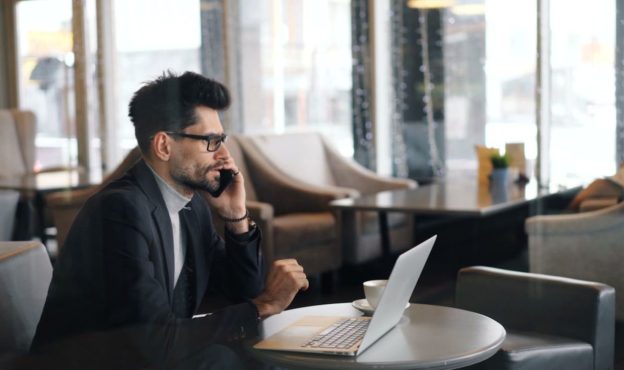 A man in a suit sitting at a table with a laptop and talking on the phone