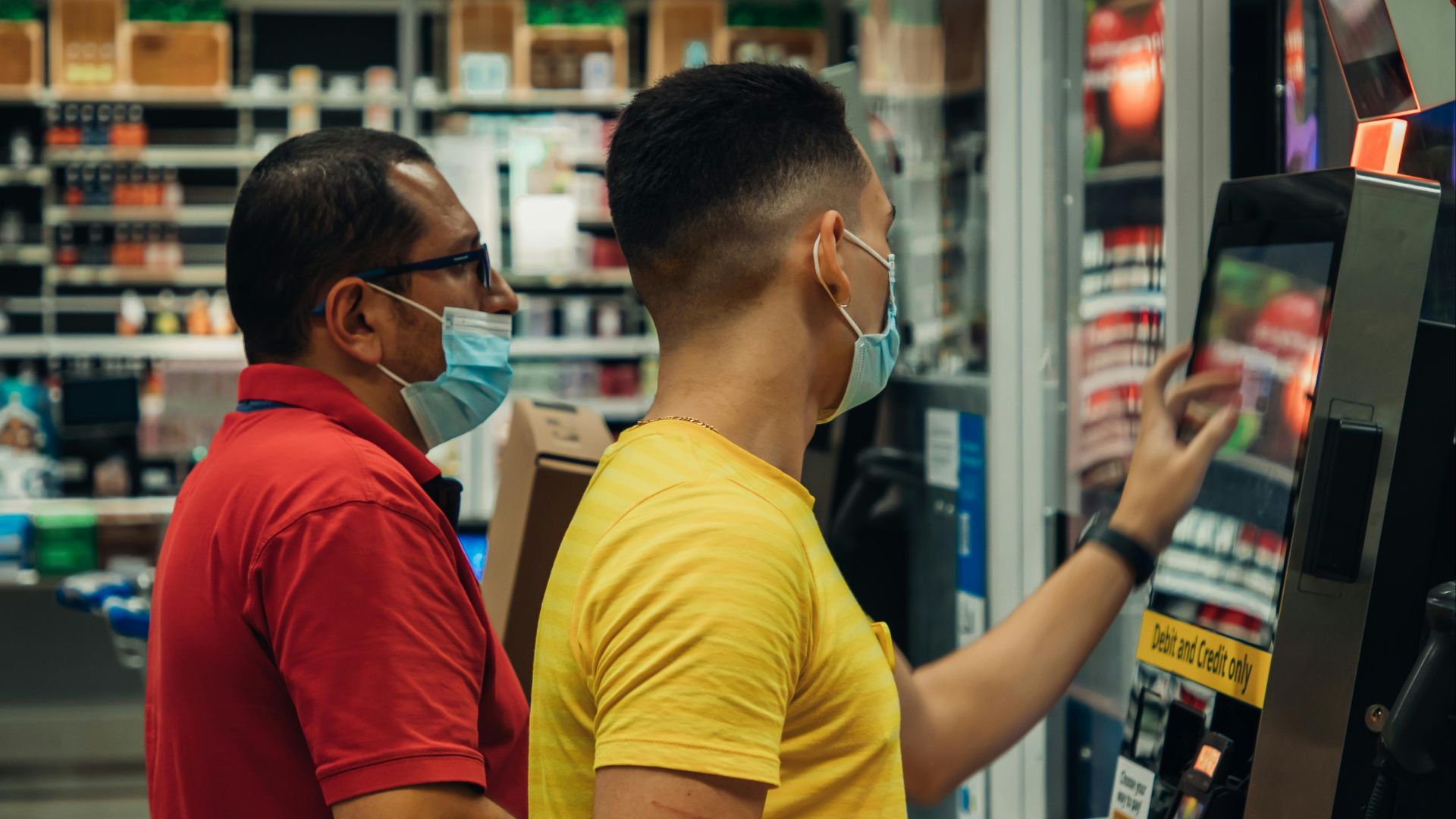 Two men wearing masks using a self-checkout machine in a grocery store.