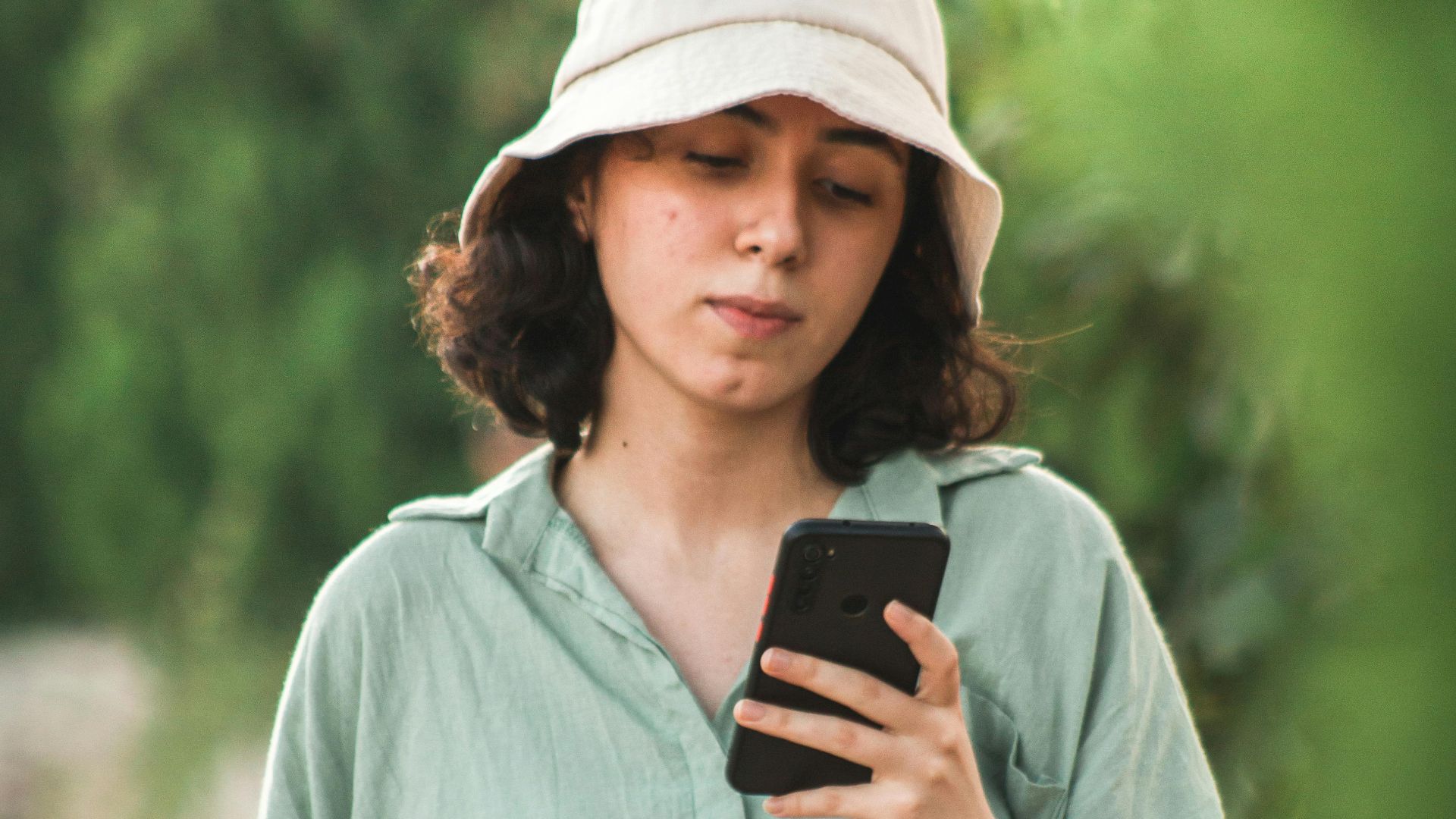 A young woman in a bucket hat uses her smartphone while walking outdoors.