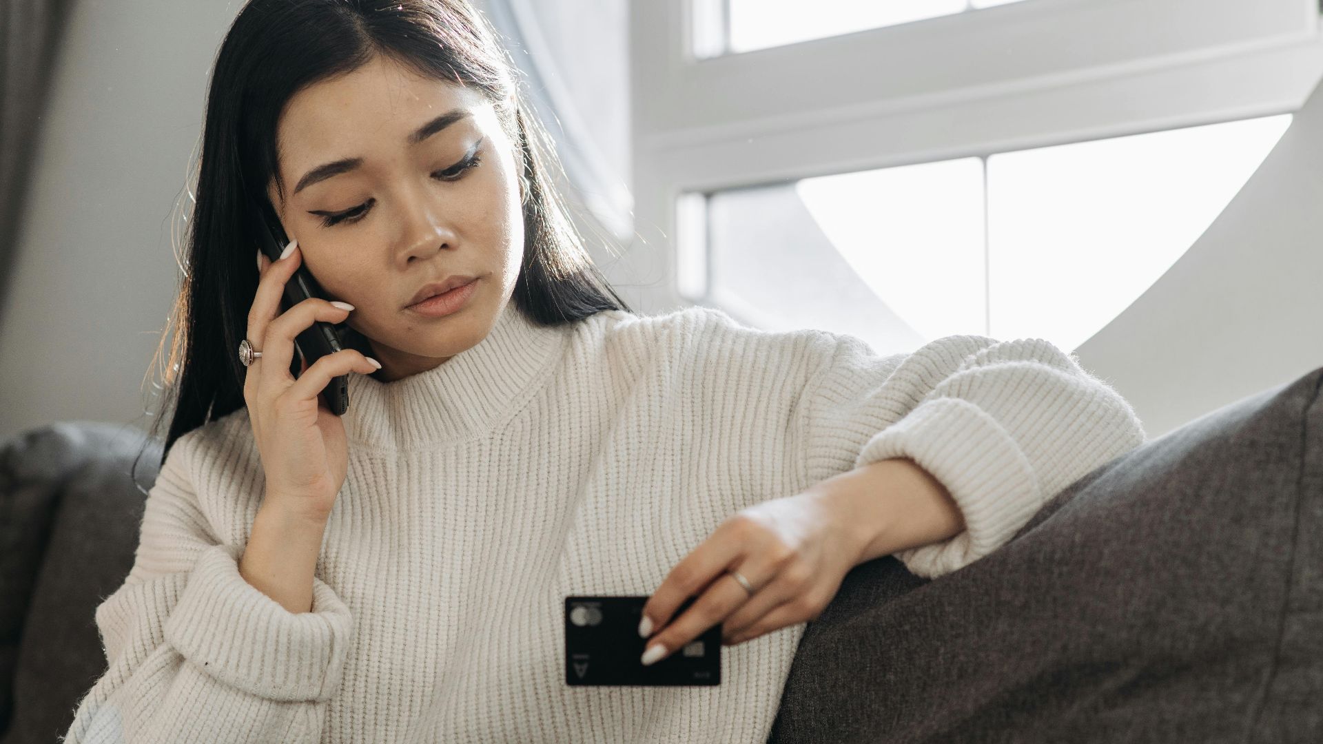 A woman sits on a sofa, multitasking with a phone and credit card, focusing on online shopping.