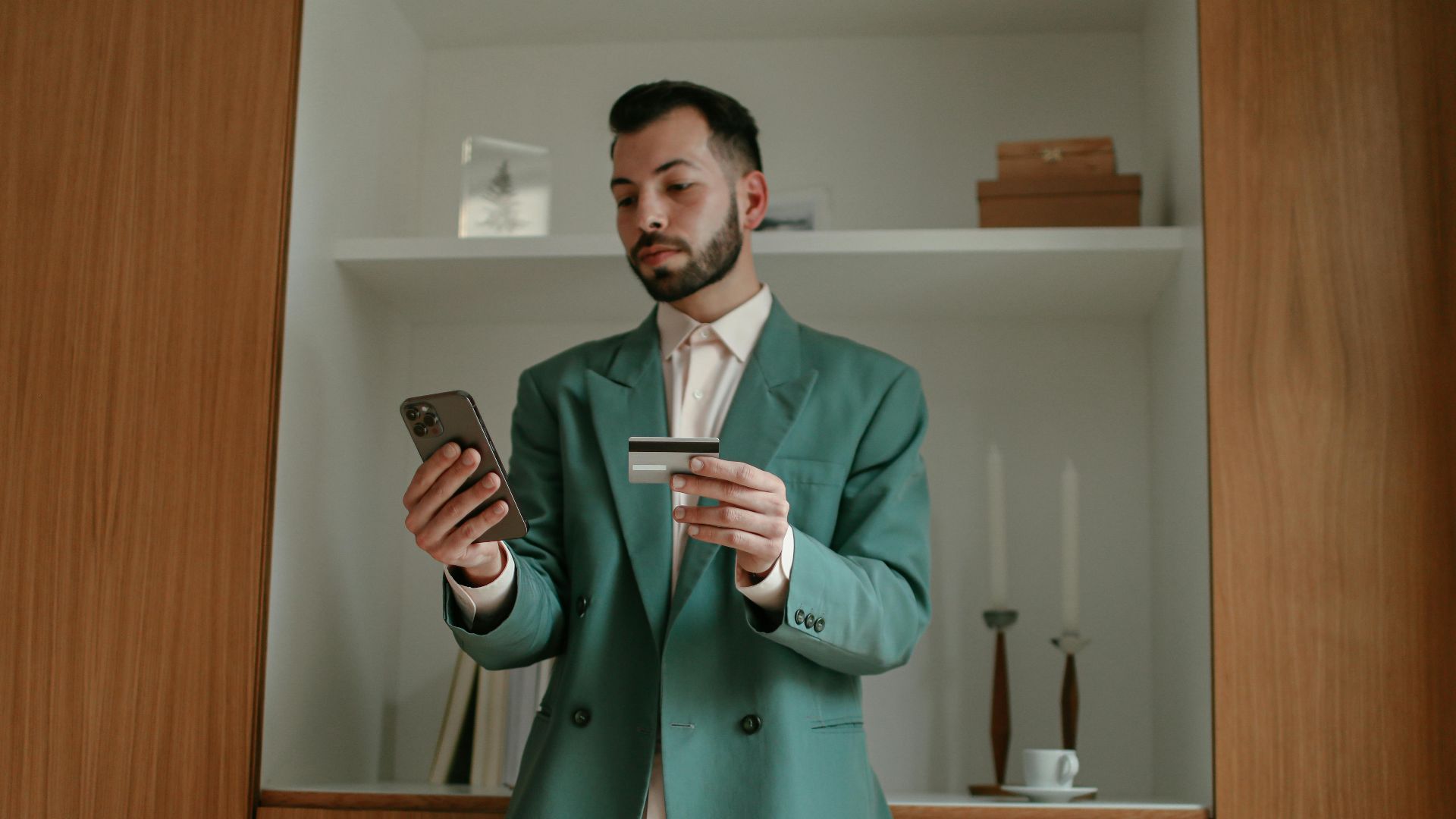 A bearded man in a green suit is using a smartphone and credit card indoors, showcasing modern technology and lifestyle.