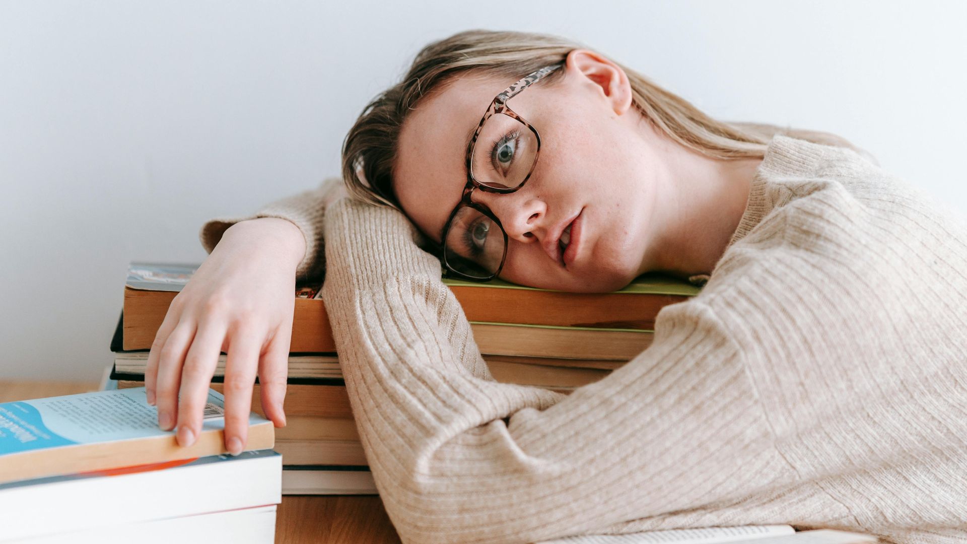 Tired female student in casual clothes lying on table with stack of books and looking away in light room in daytime