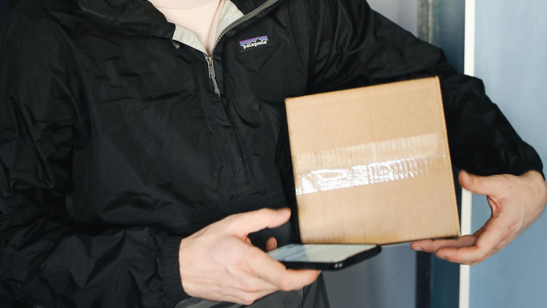 A delivery man holds a package and uses his smartphone inside a building.
