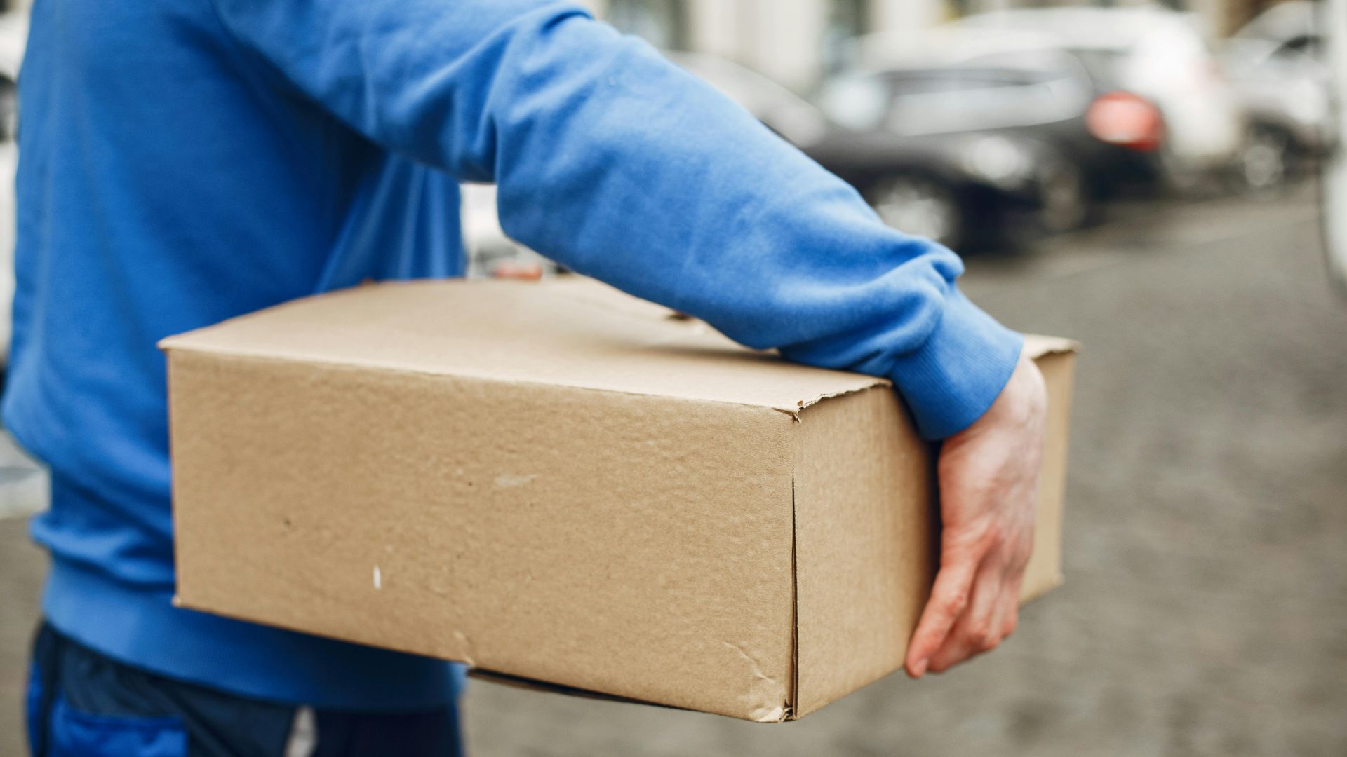A courier carrying a cardboard box outdoors on a city street, offering delivery service.