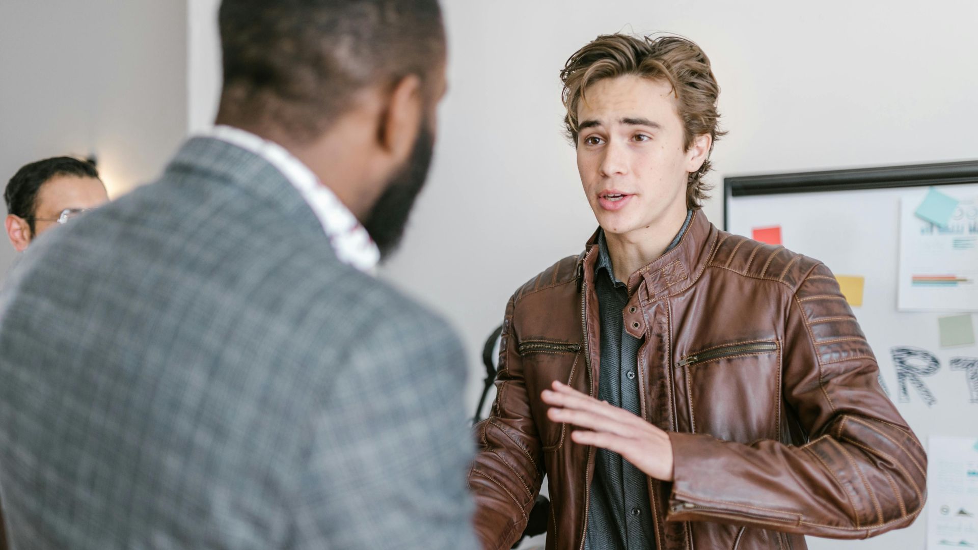 A young man in a leather jacket talking with colleagues during a business meeting indoors.