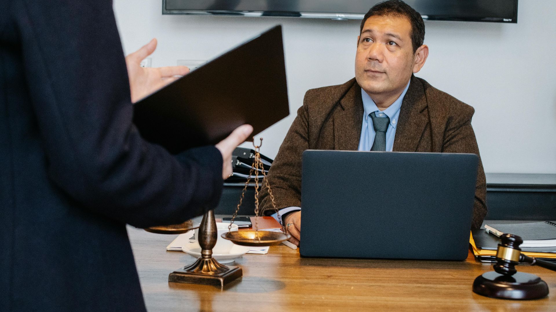 Crop unrecognizable employee representing new case details to concentrated middle aged ethnic lawyer sitting at table with laptop gavel and justice scales